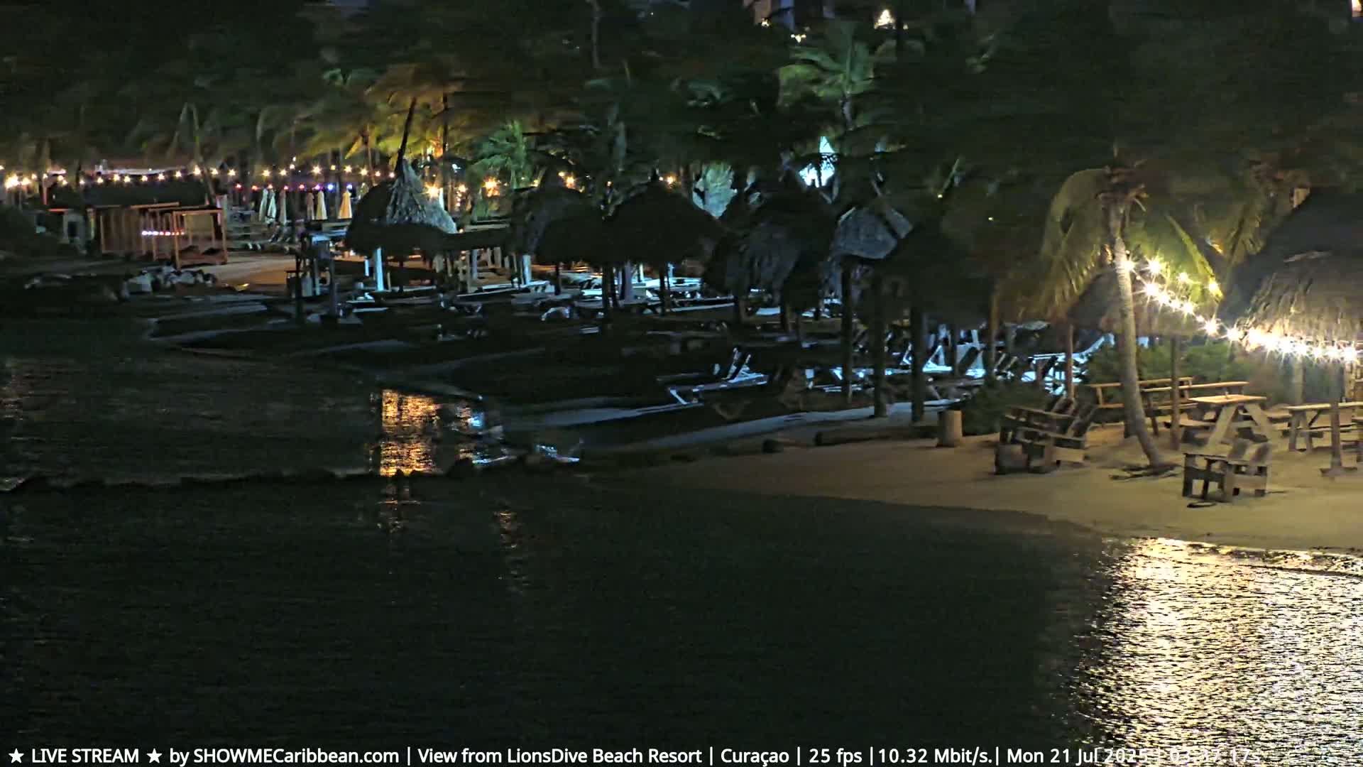 A nighttime beach scene shows numerous thatched-roof structures and picnic tables illuminated by string lights, alongside calm water reflecting the lights.