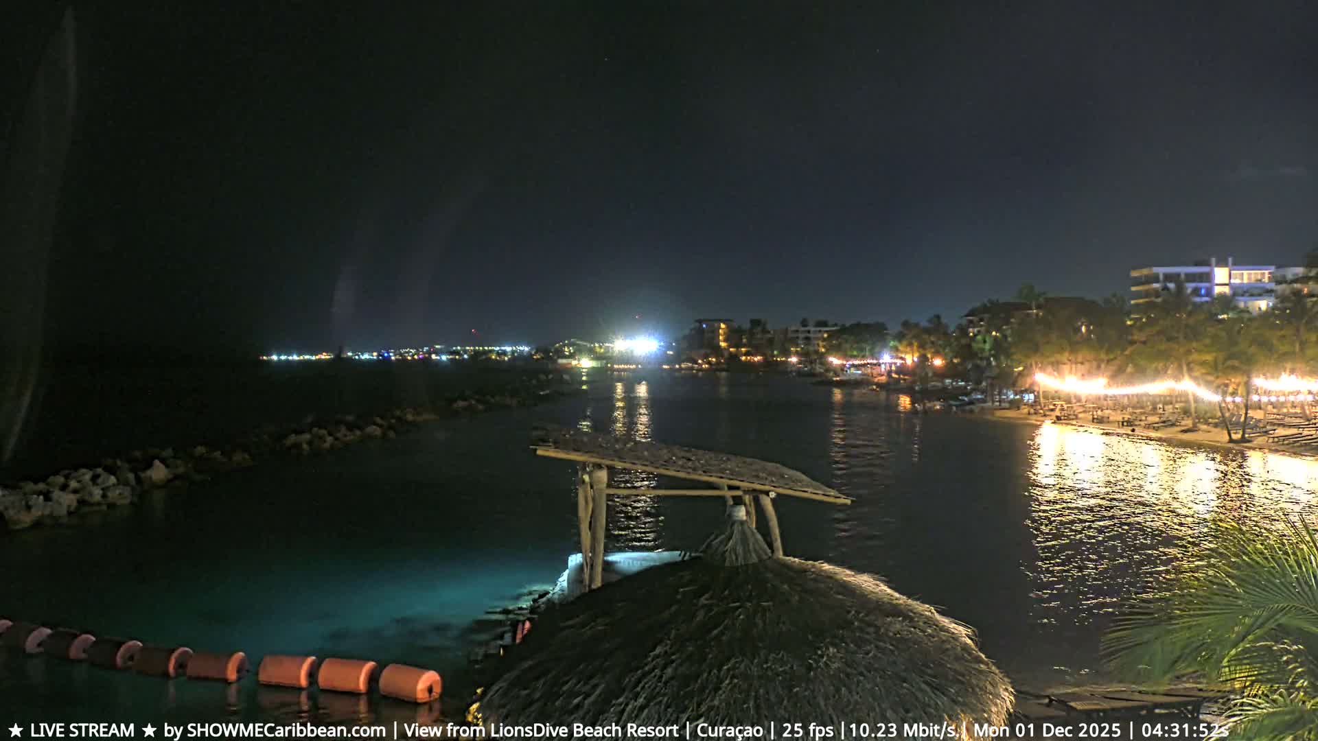 The image shows a serene nighttime view of a coastal resort, featuring a brightly lit beach with palm trees and buildings reflecting on calm waters, a thatched hut in the foreground, and a distant illuminated cityscape under a clear sky.