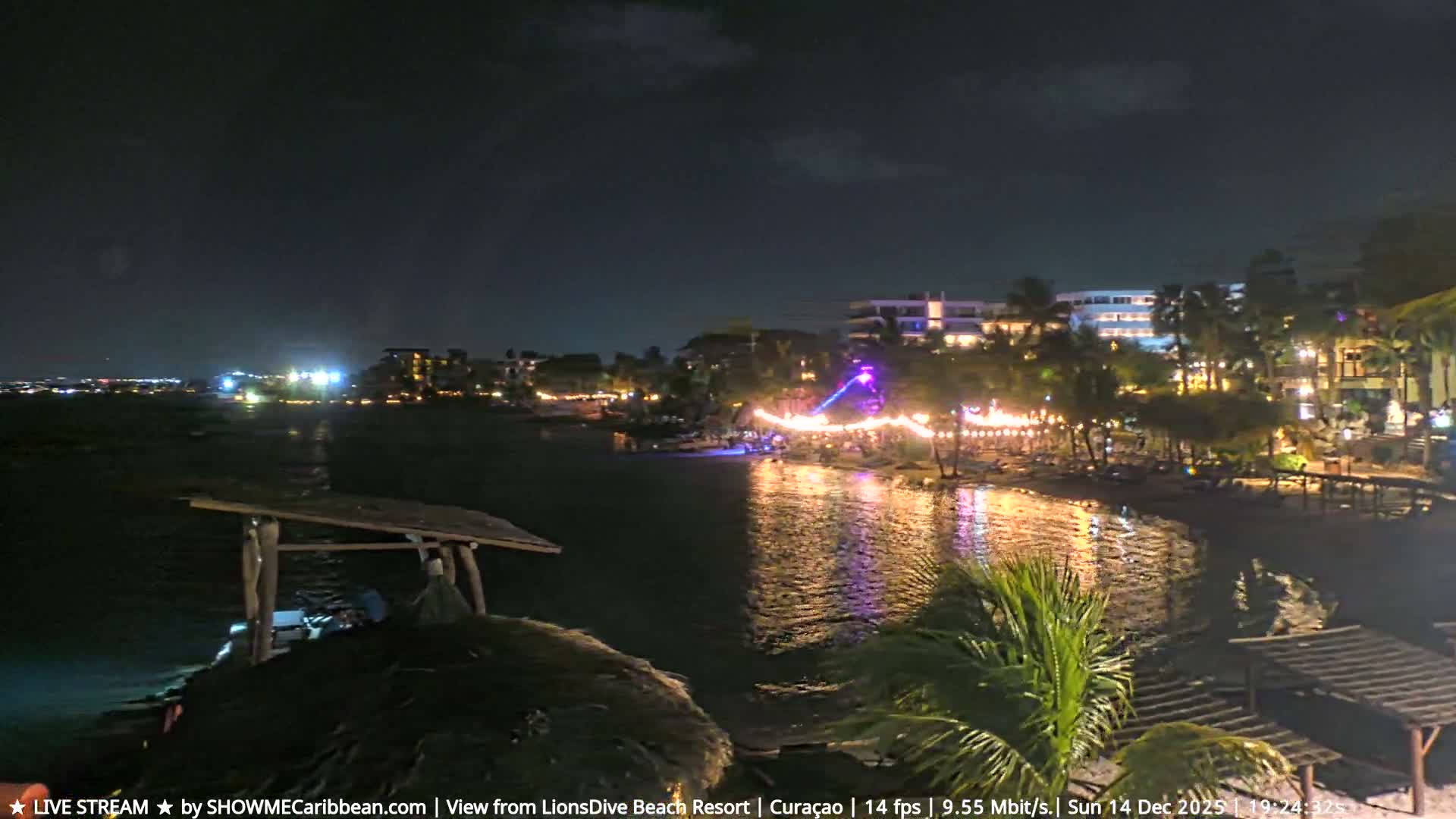 The image shows a resort beach or waterfront at night, illuminated by string lights and colorful uplighting on the sand where picnic tables and lounge chairs are set up, with the lights reflecting on the calm water under clear skies.