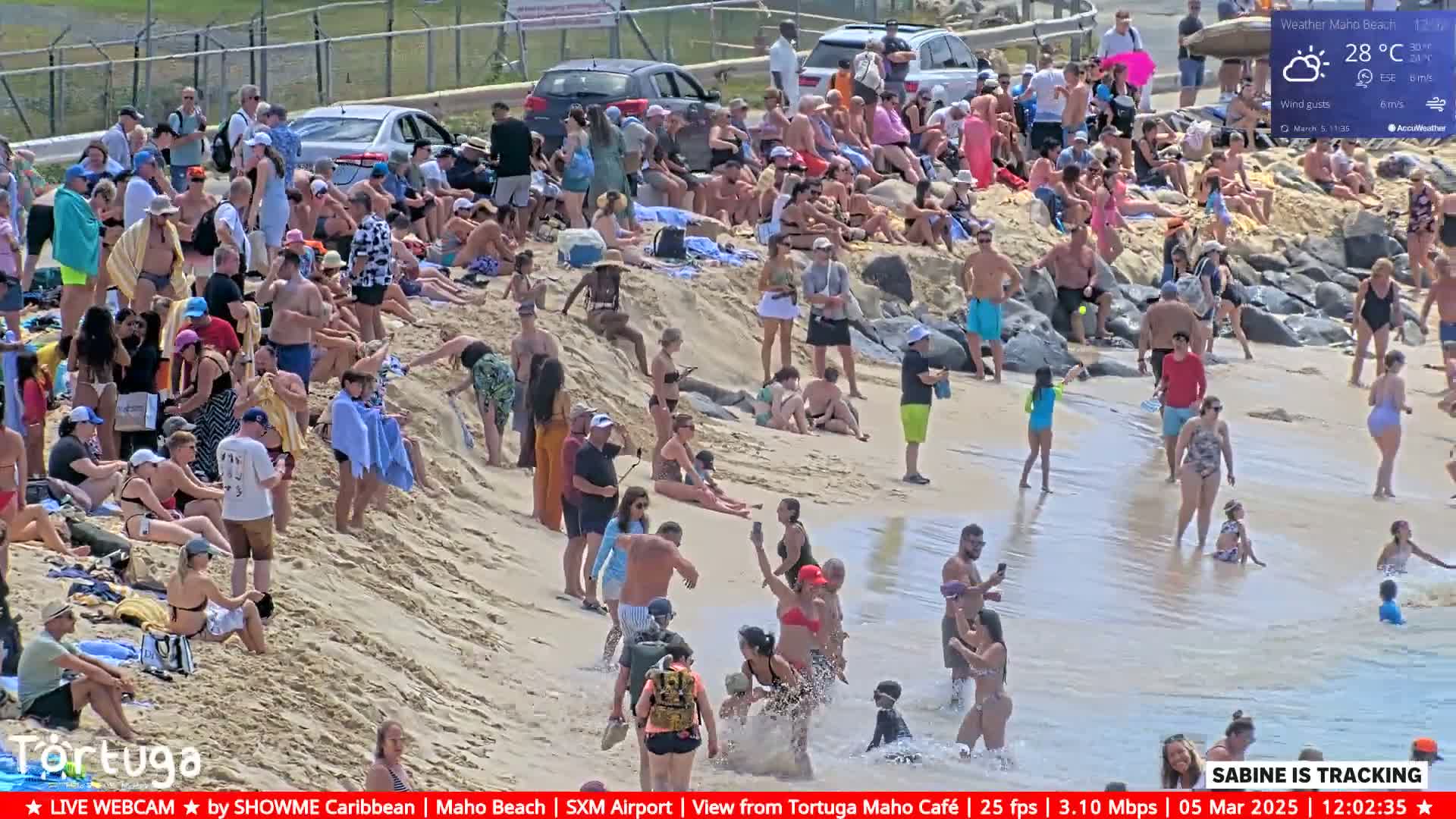 A large crowd of people are gathered on a sandy beach near the water under partly cloudy skies; some people are in the water.