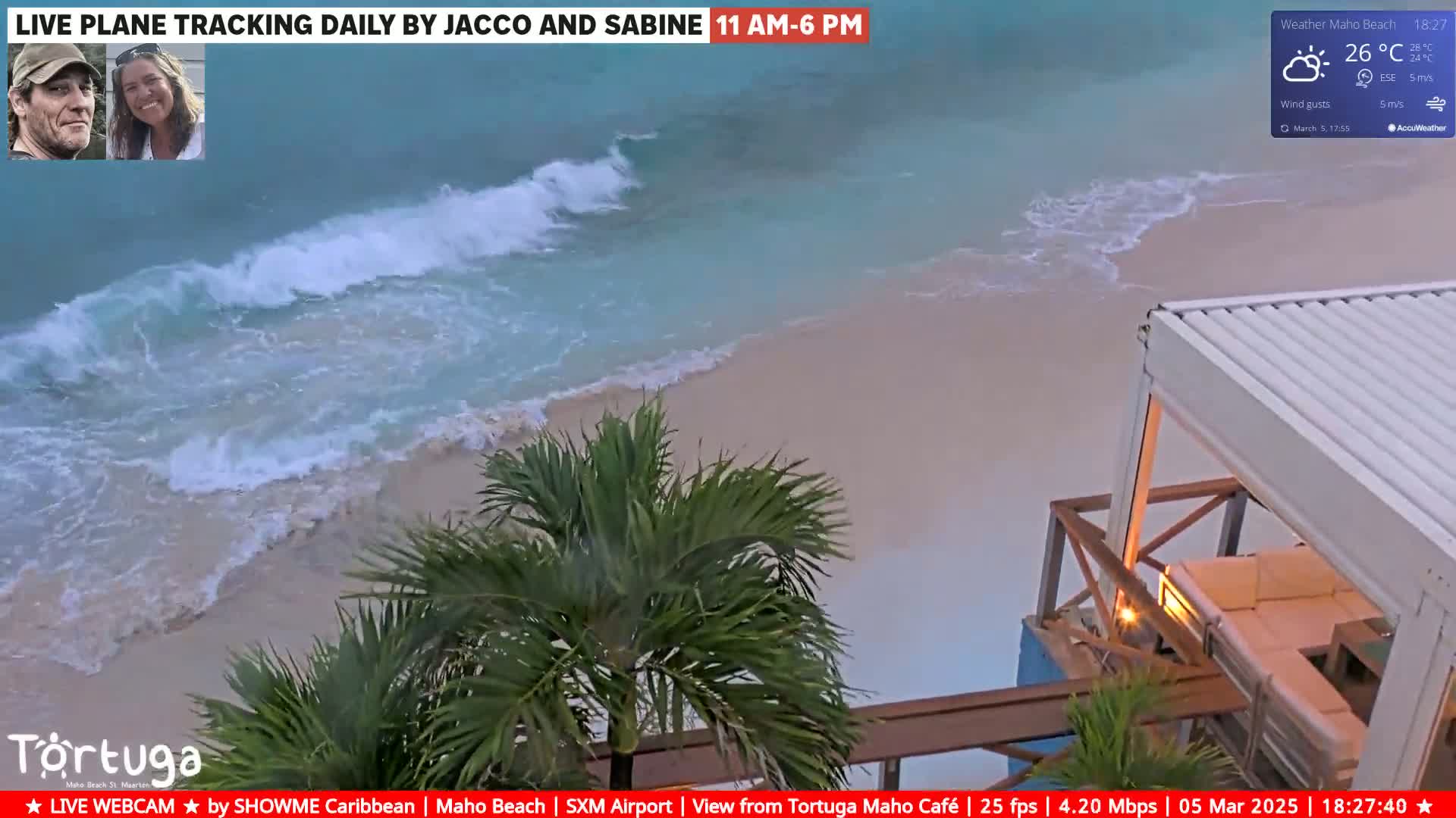 Ocean waves lap a sandy beach near a partially covered outdoor seating area under partly cloudy skies.