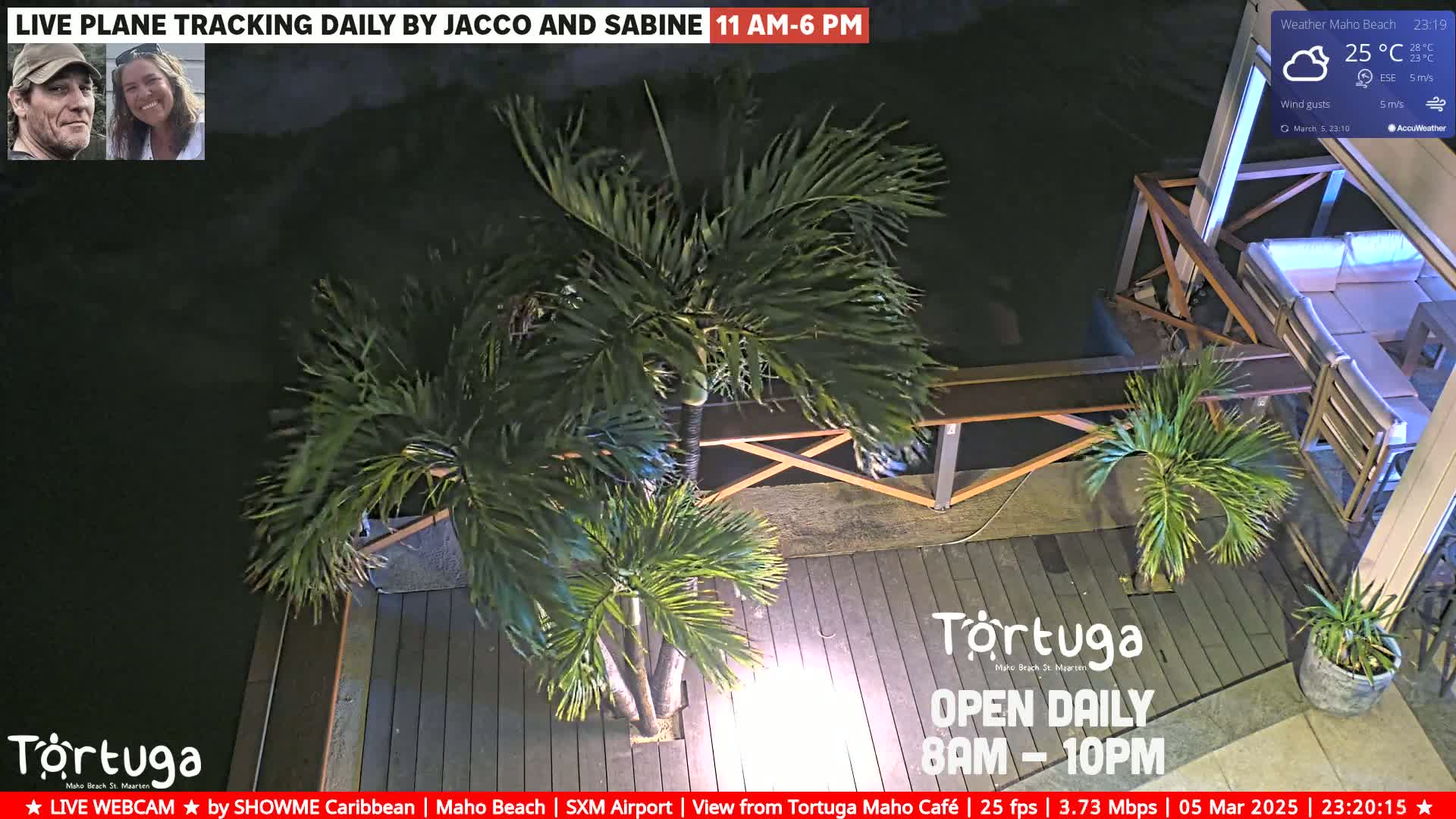 A nighttime outdoor scene shows a wooden deck with potted palm trees, outdoor furniture, and a dark body of water beyond.
