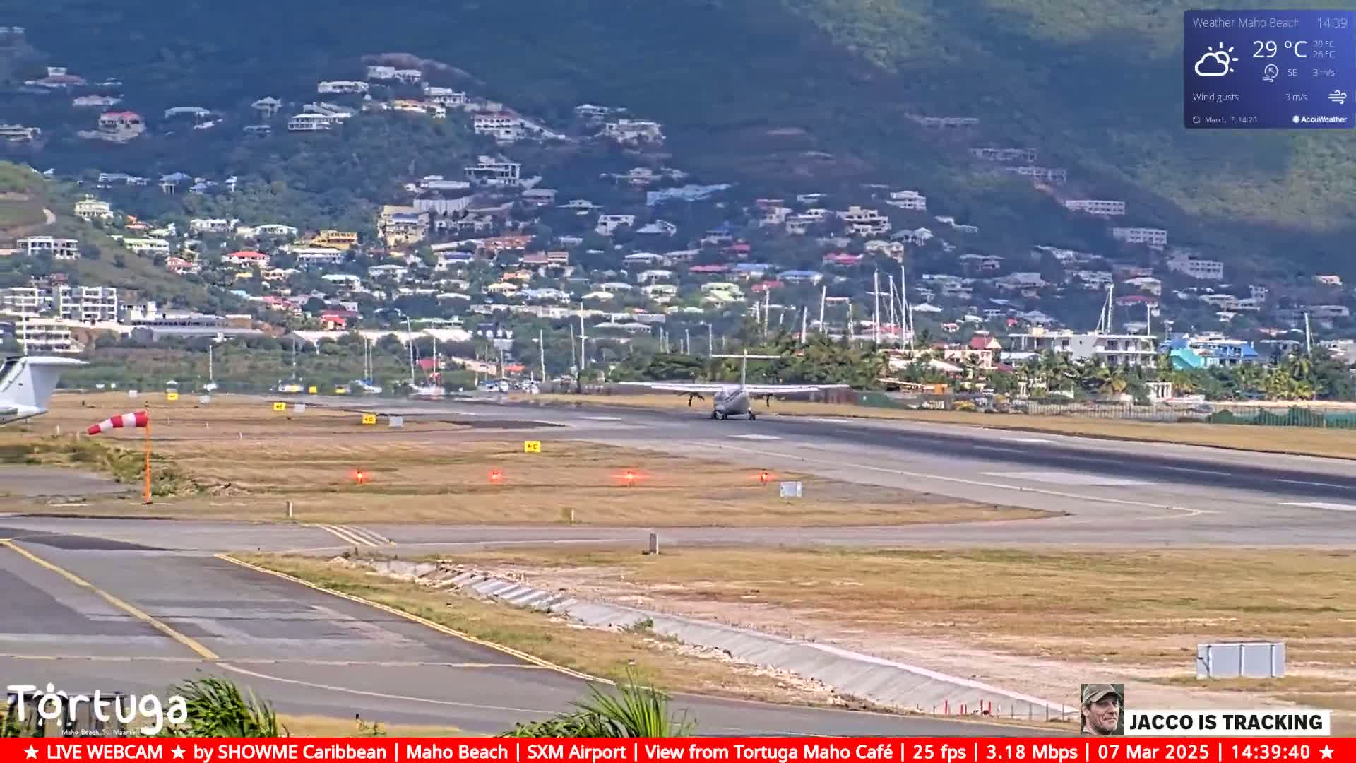 A small airplane taxis on a runway near a hillside town under partly cloudy skies.