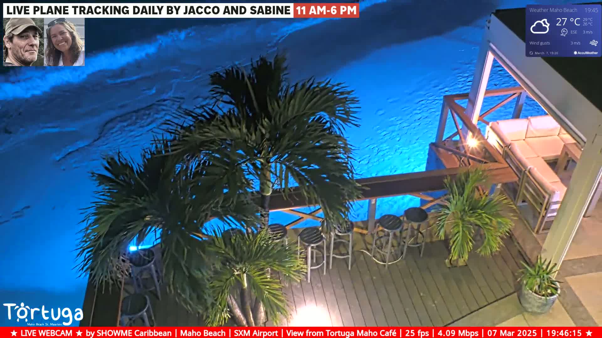 A nighttime view of an outdoor patio area with a seating arrangement overlooking a brightly lit blue body of water, under a partly cloudy sky.