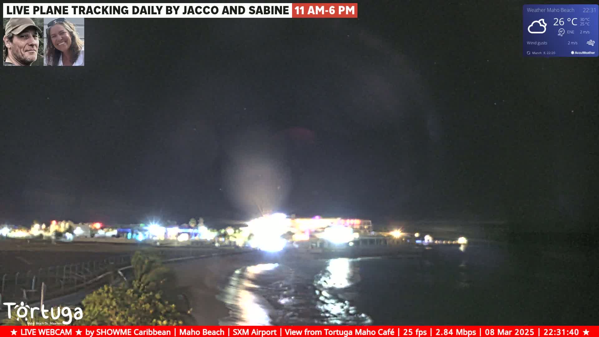 A nighttime beach scene shows a shoreline with lights from buildings and a calm ocean under a partly cloudy sky.