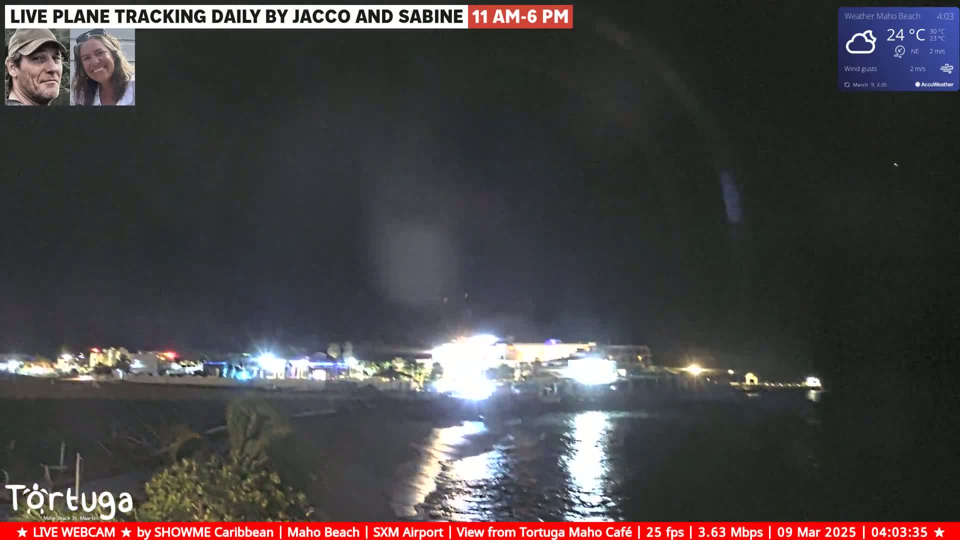 A nighttime coastal scene with partly cloudy skies shows a dark body of water lapping at a shoreline with illuminated buildings in the distance.