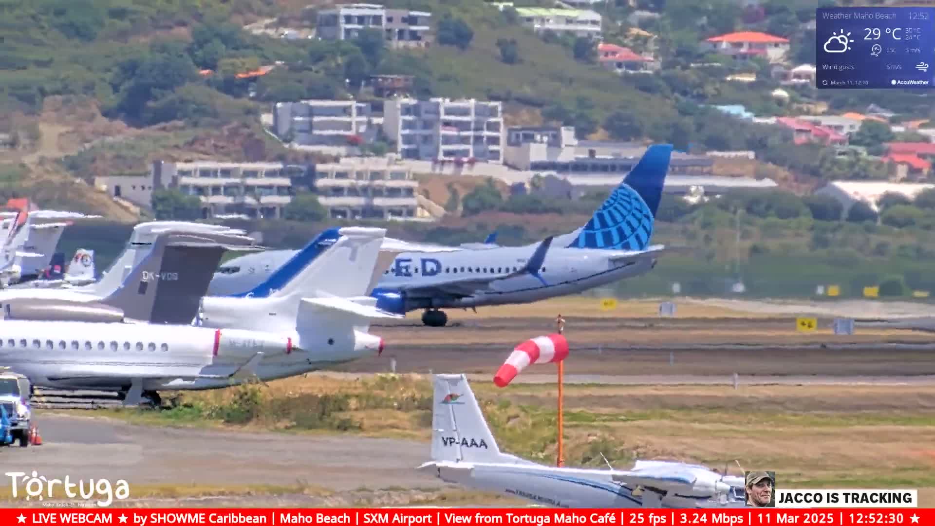 A commercial jet is landing on an airport runway near several smaller planes and a building, under partly sunny skies.