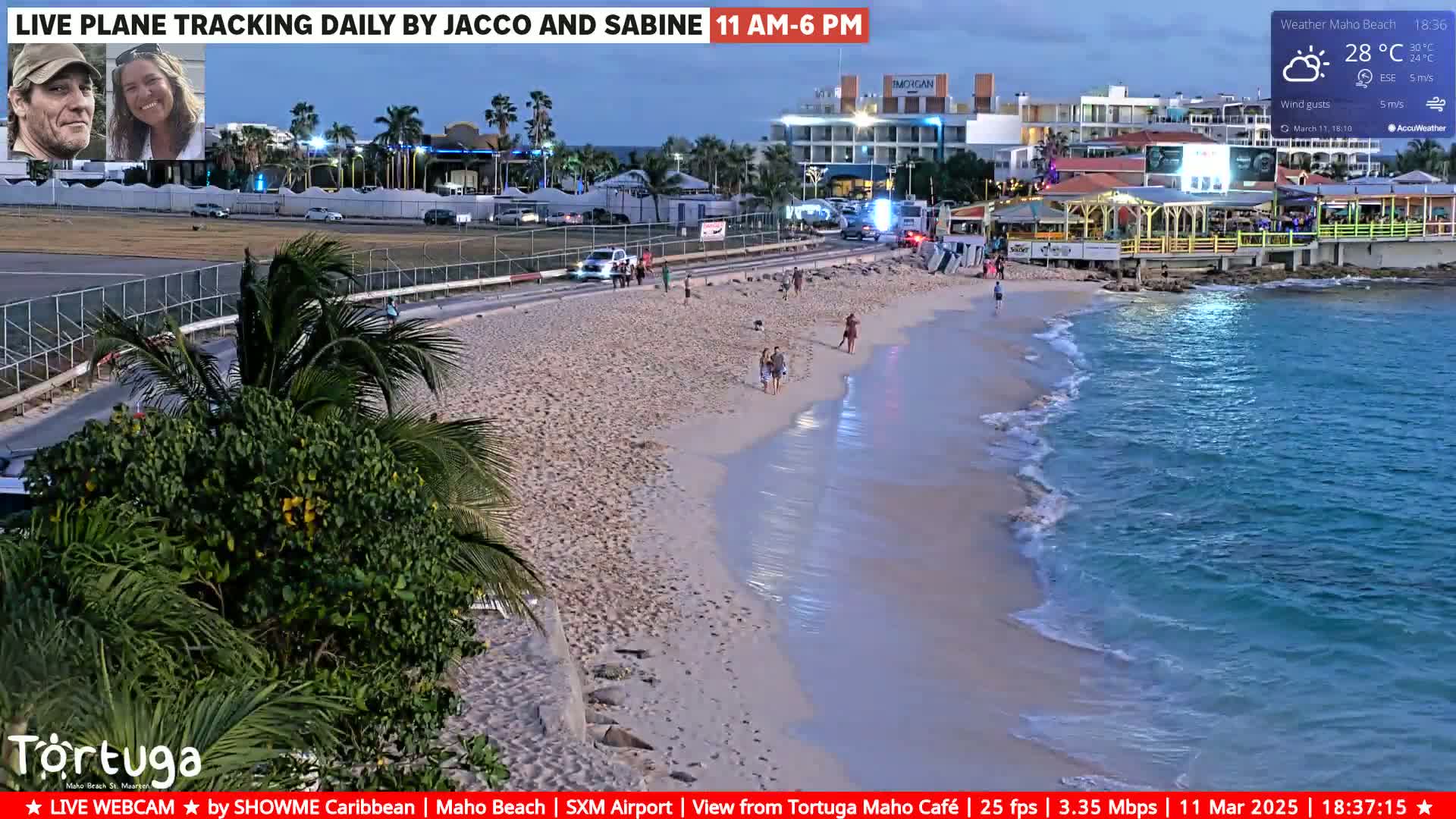 A sandy beach with calm, clear water laps the shore under a partly cloudy twilight sky; several people are strolling along the beach near a road and buildings.