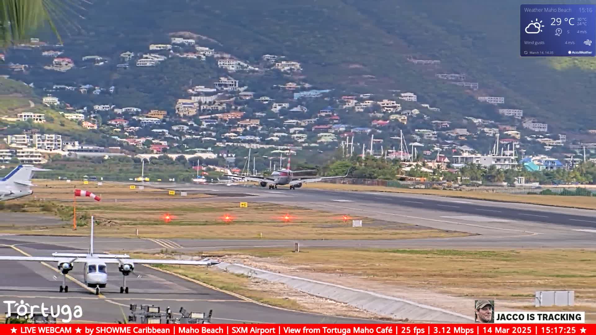 A passenger plane lands on a runway at an airport, with another plane taxiing nearby, under partly sunny skies and a light wind, overlooking a hillside town.