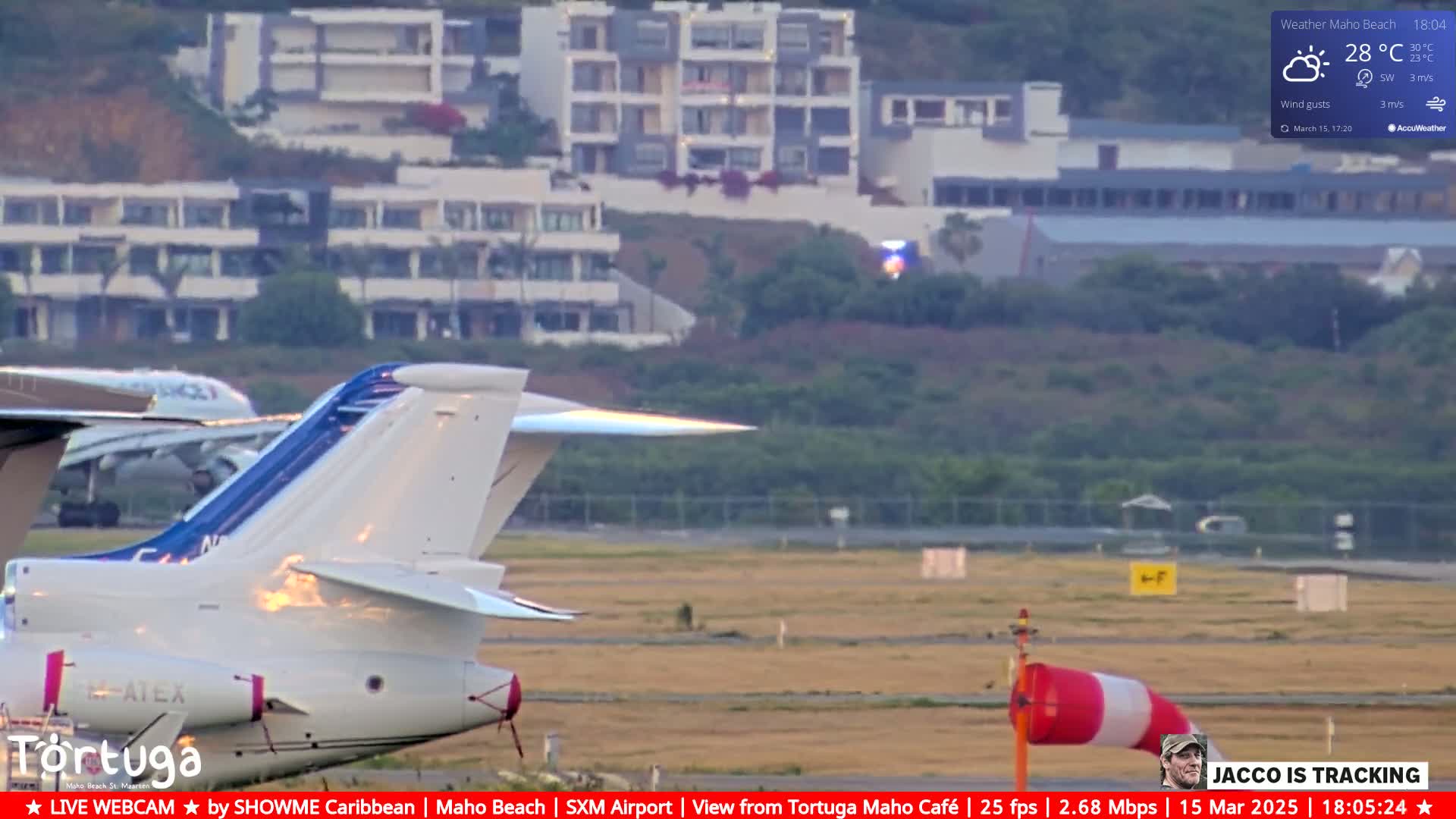 A white airplane's tail and part of its body are visible on an airfield, with another airplane in the background and a multi-story building in the distance under partly sunny skies.