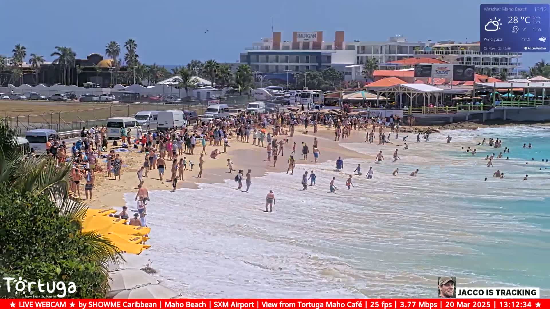A crowded beach with people swimming and sunbathing under sunny skies, with buildings and parked cars visible in the background.