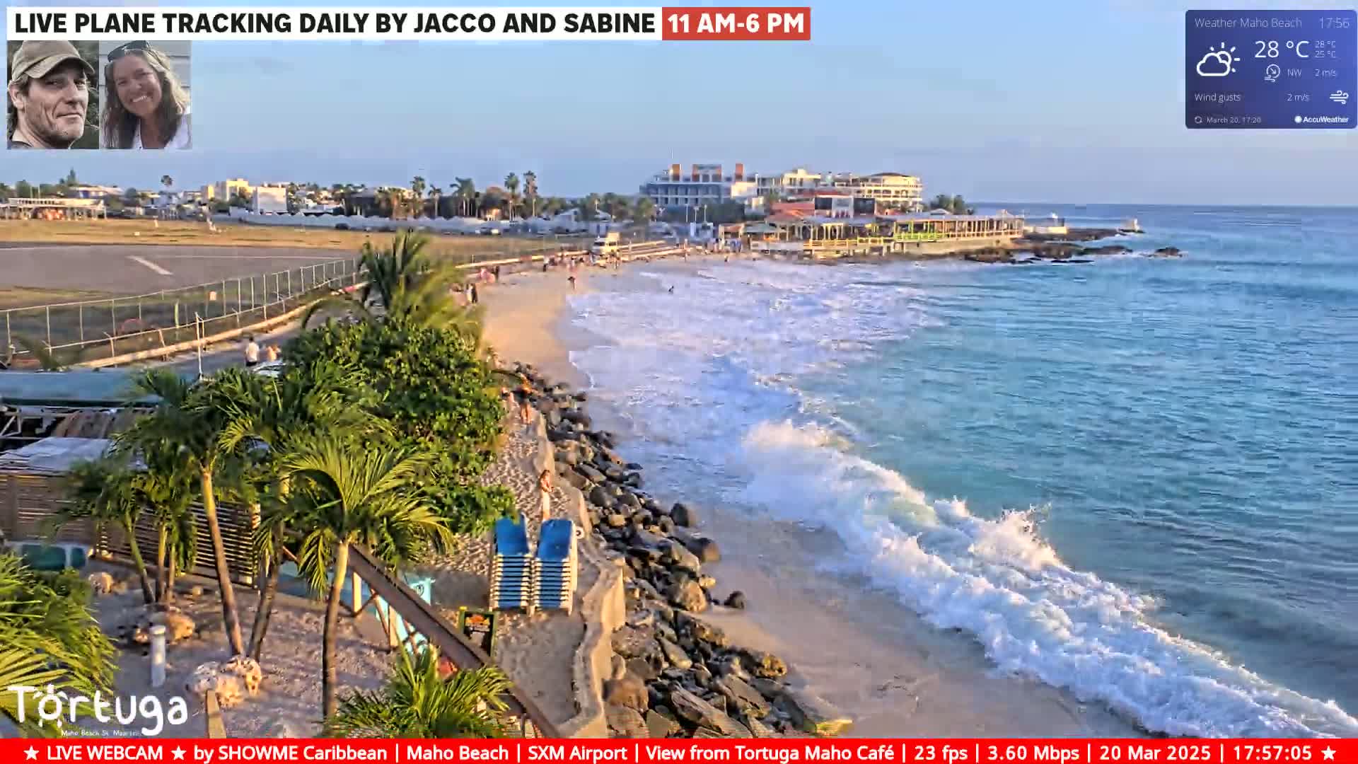 A beach scene shows a sandy shore with gentle waves lapping, a rocky shoreline, some buildings in the background, and a few people on the beach under a partly sunny sky.