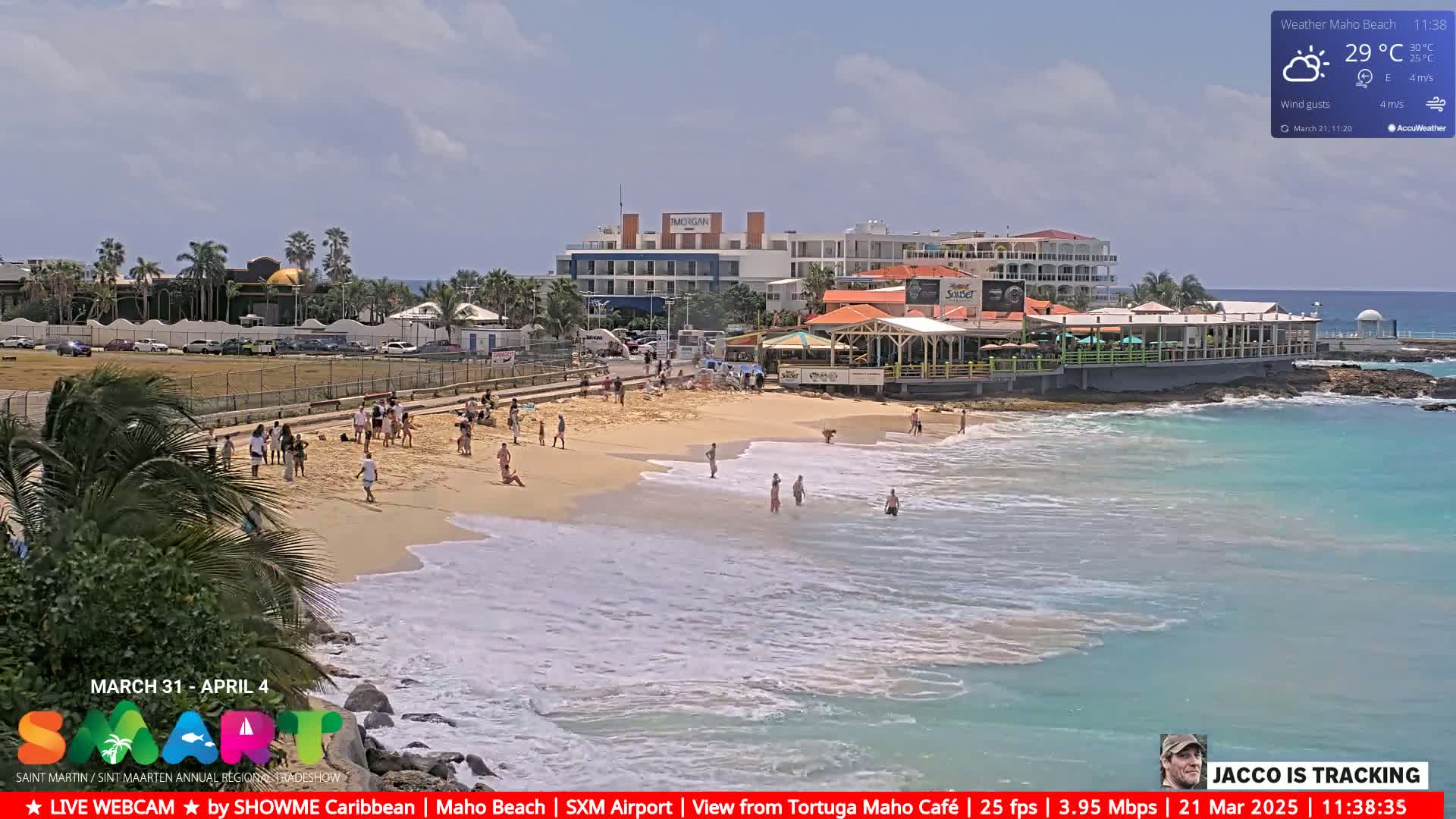 A partly sunny beach scene shows people relaxing and swimming in turquoise water, with buildings and a restaurant in the background.