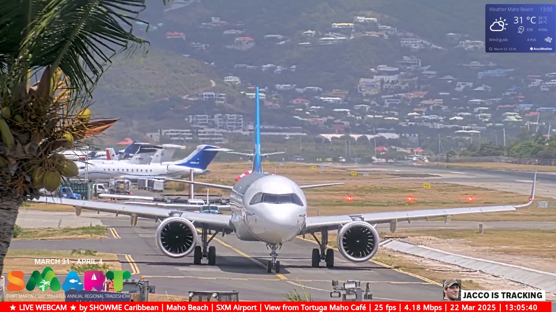 A silver airplane taxis on an airport runway near a hill dotted with houses under partly sunny skies.