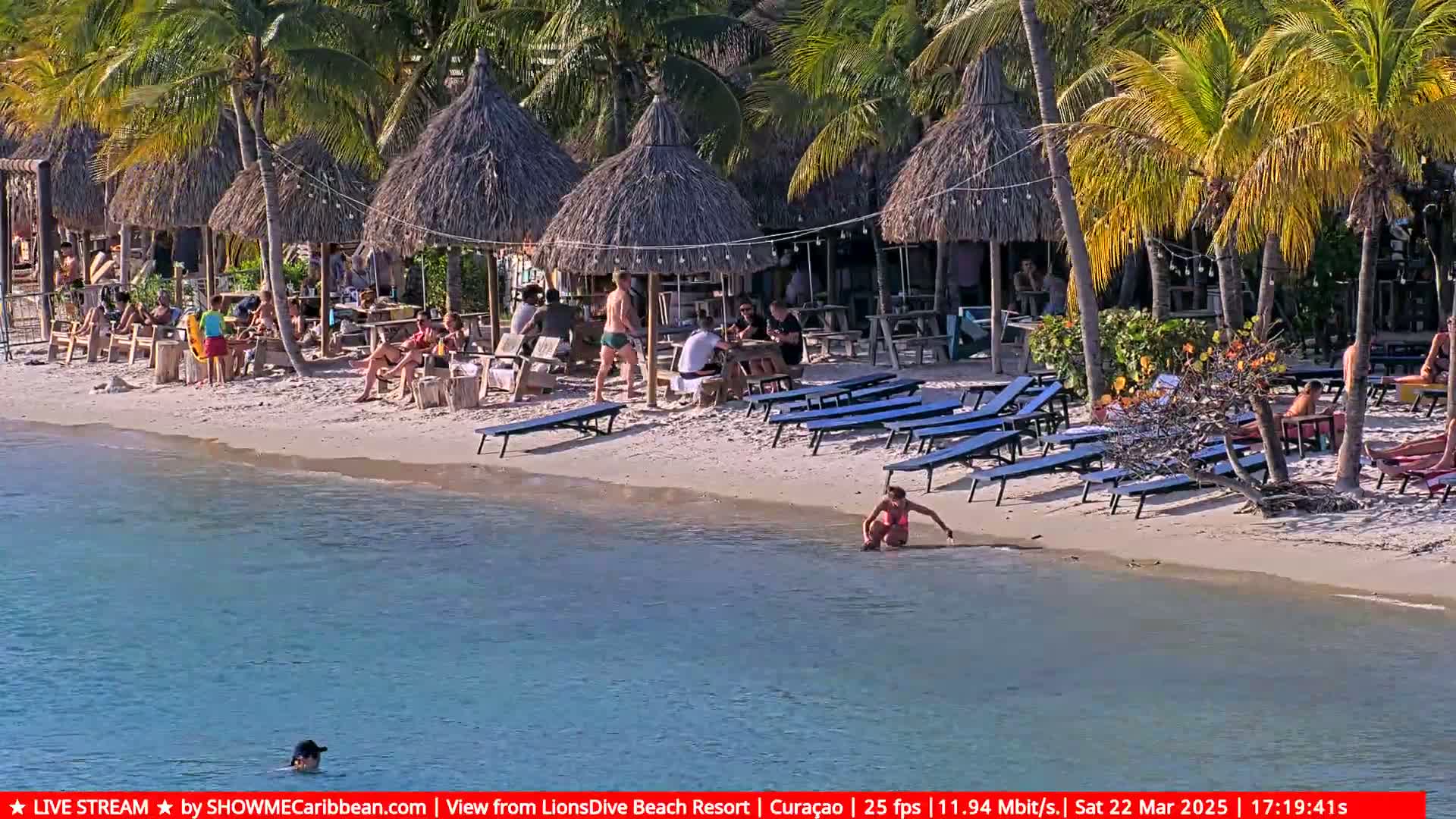 A sunny beach scene shows numerous people relaxing under thatched-roof huts and lounge chairs near calm, shallow water.