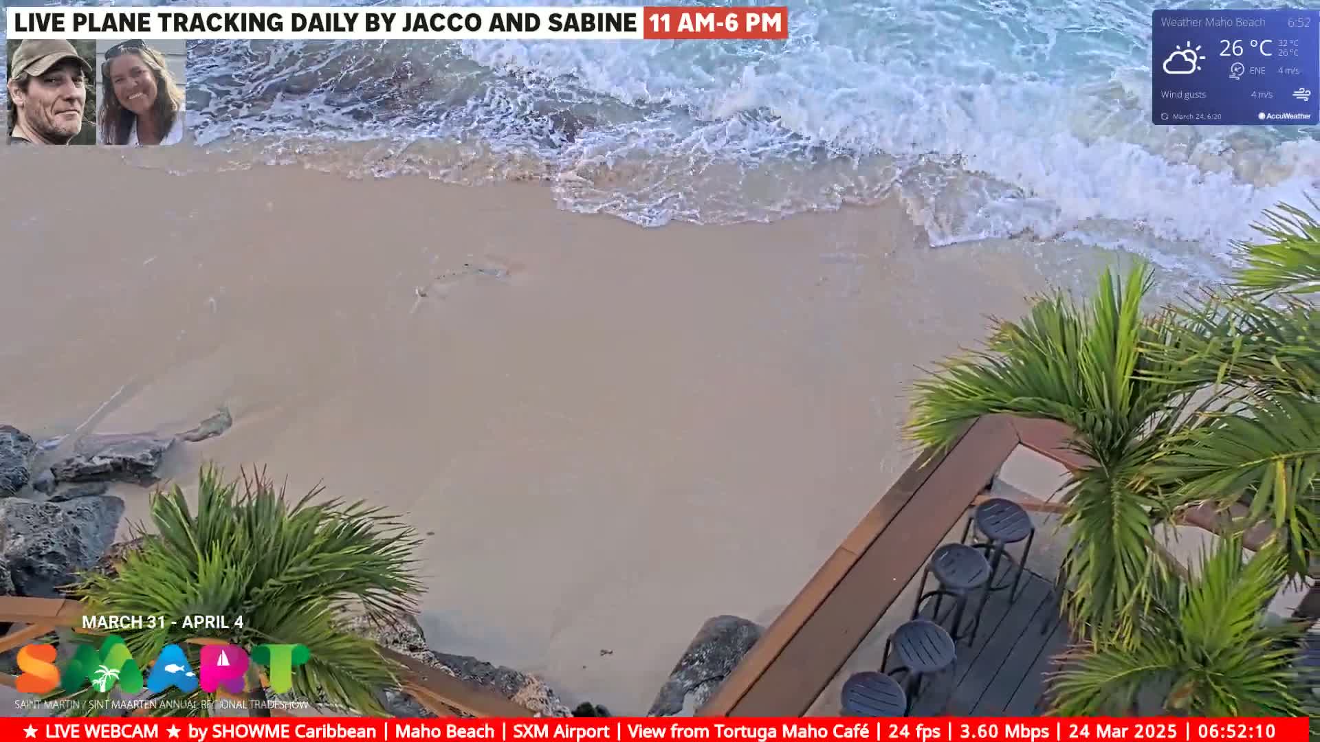 A sandy beach with gentle waves laps the shore under partly sunny skies, viewed from an elevated perspective showing palm trees and outdoor cafe seating.