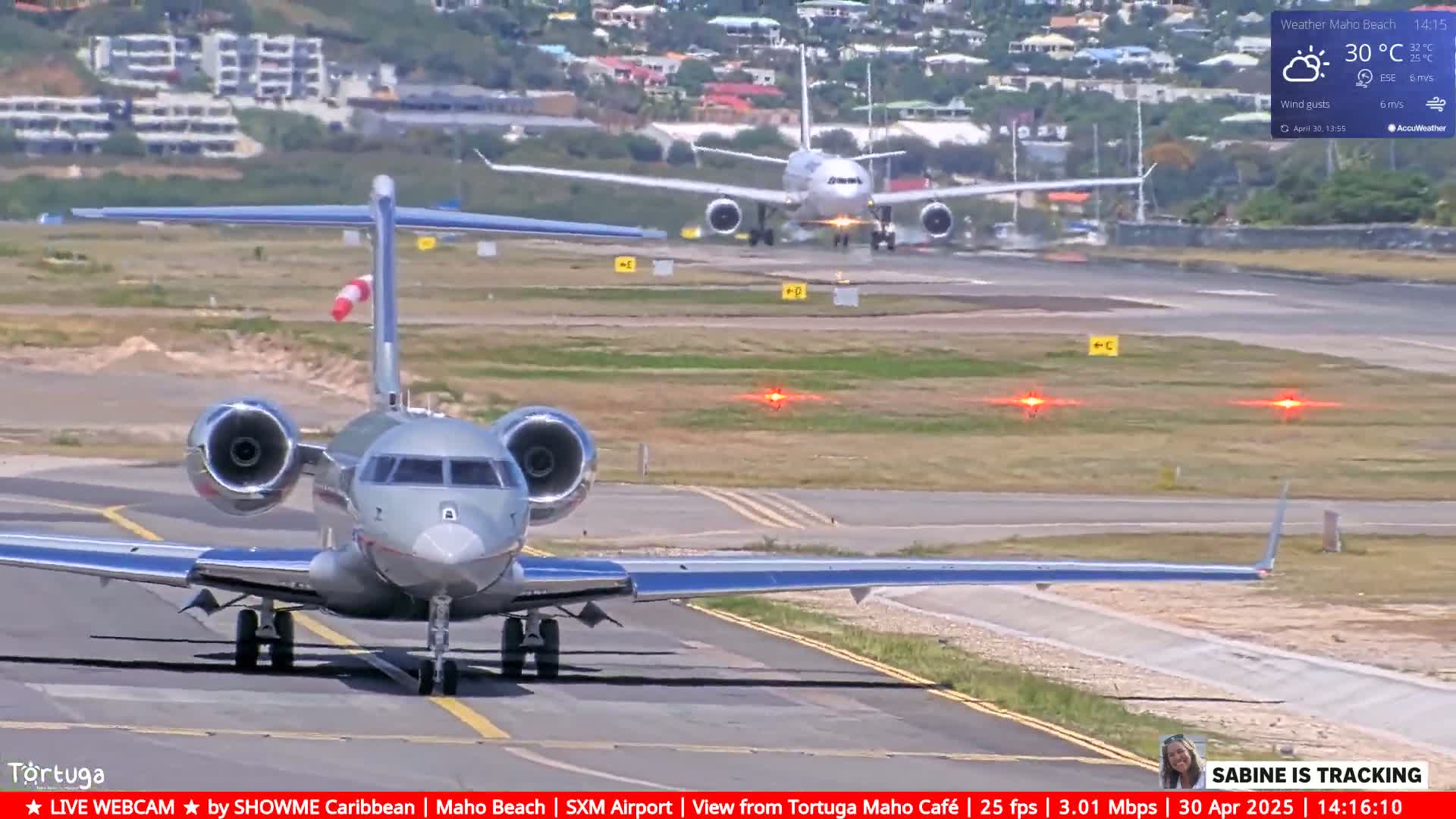 A private jet is taxiing on an airport runway in sunny weather, with a larger passenger plane visible in the background.
