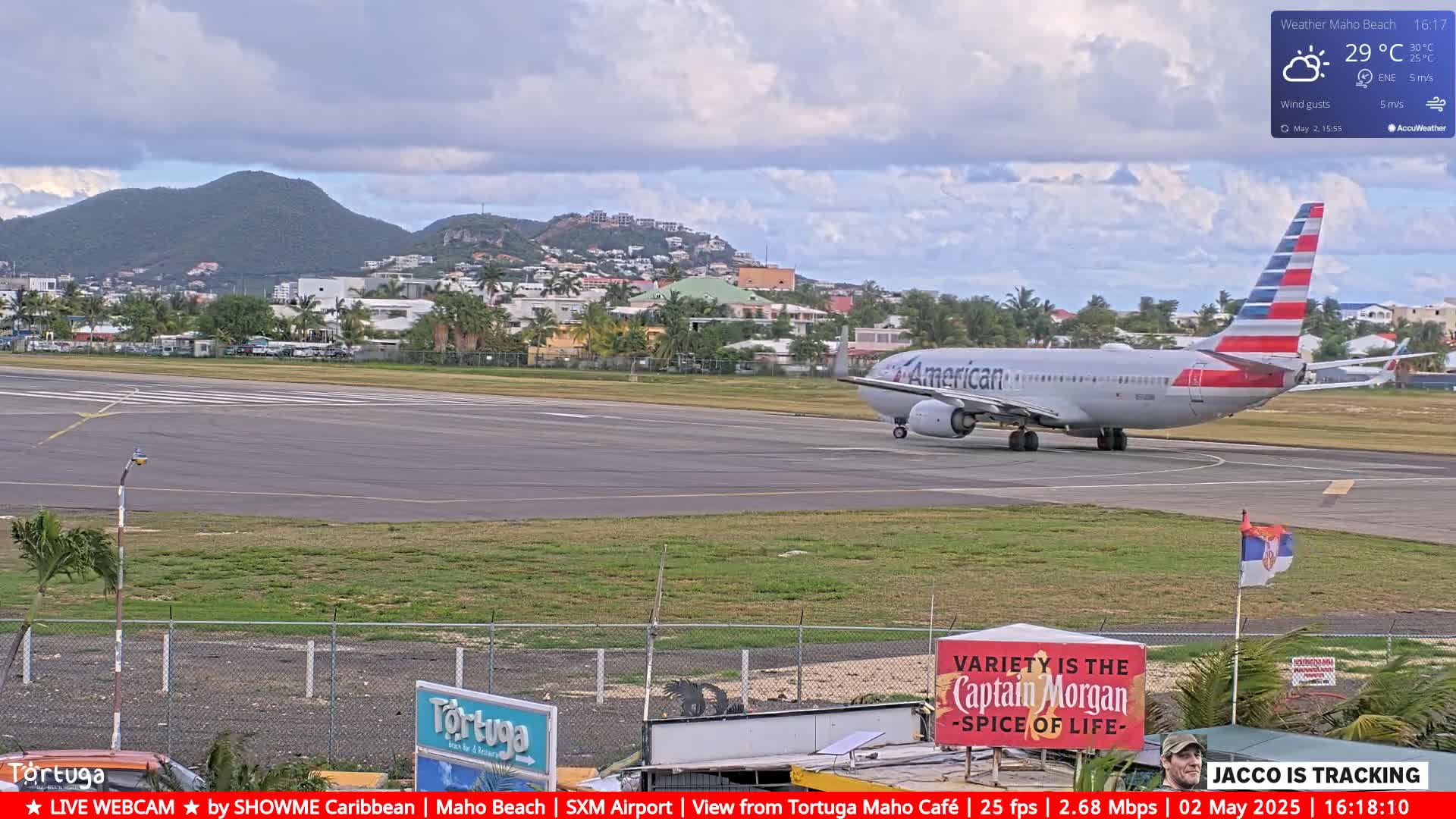 An American Airlines jet taxis on an airport runway near a town nestled against a mountainous backdrop under a partly cloudy sky.