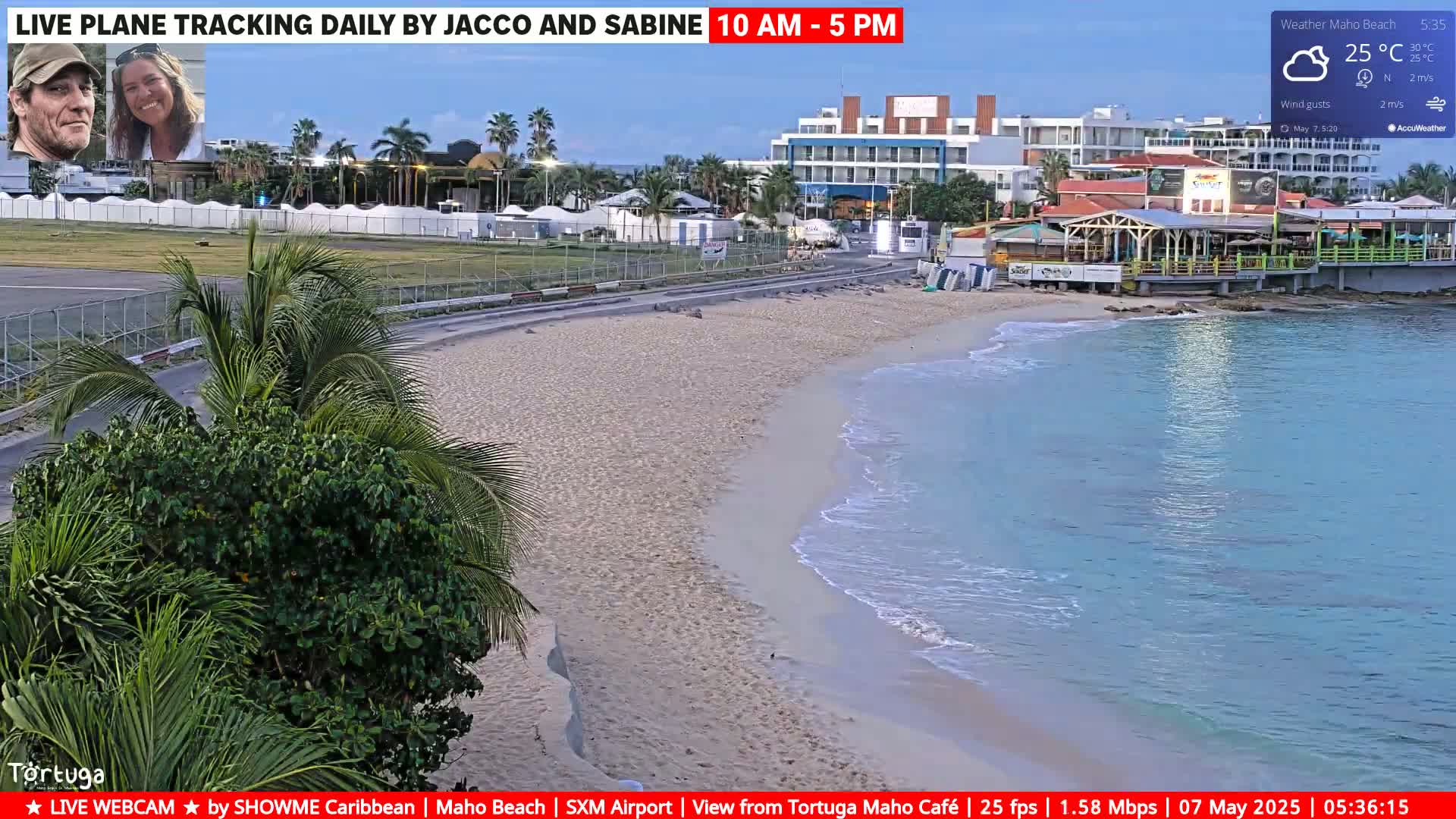 A sandy beach with calm, light blue water laps the shore near buildings and palm trees under a partly cloudy sky.