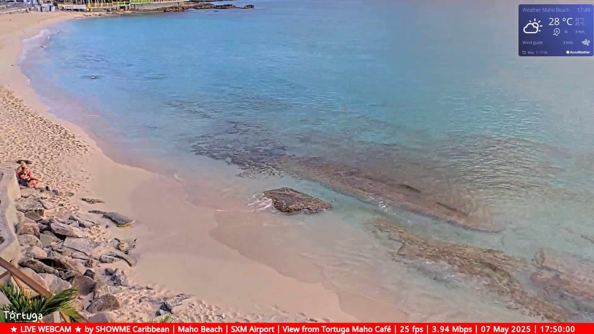 A sandy beach with clear, shallow turquoise water laps gently at the shore, where a couple sits; rocks are visible along the edge of the beach and in the water.