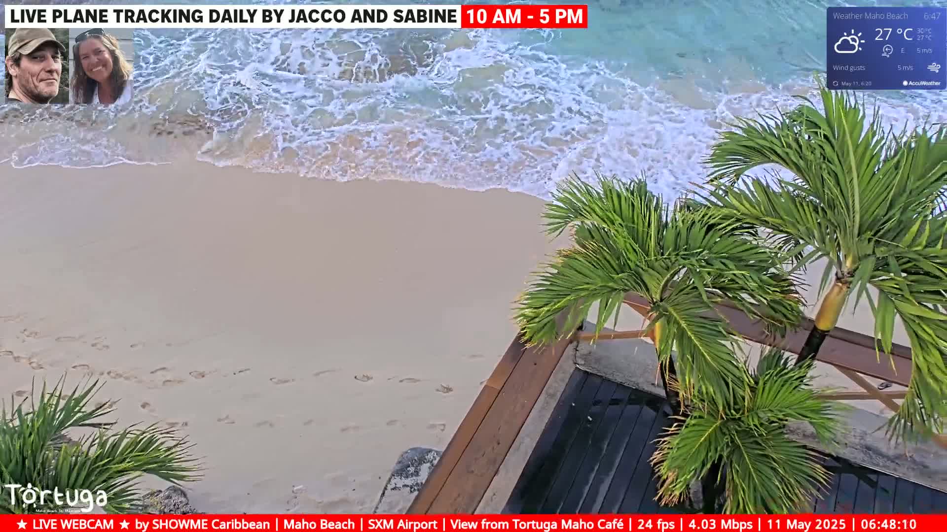 A sandy beach with footprints and gentle waves under a partly cloudy sky, partially viewed from above a wooden deck and palm trees.