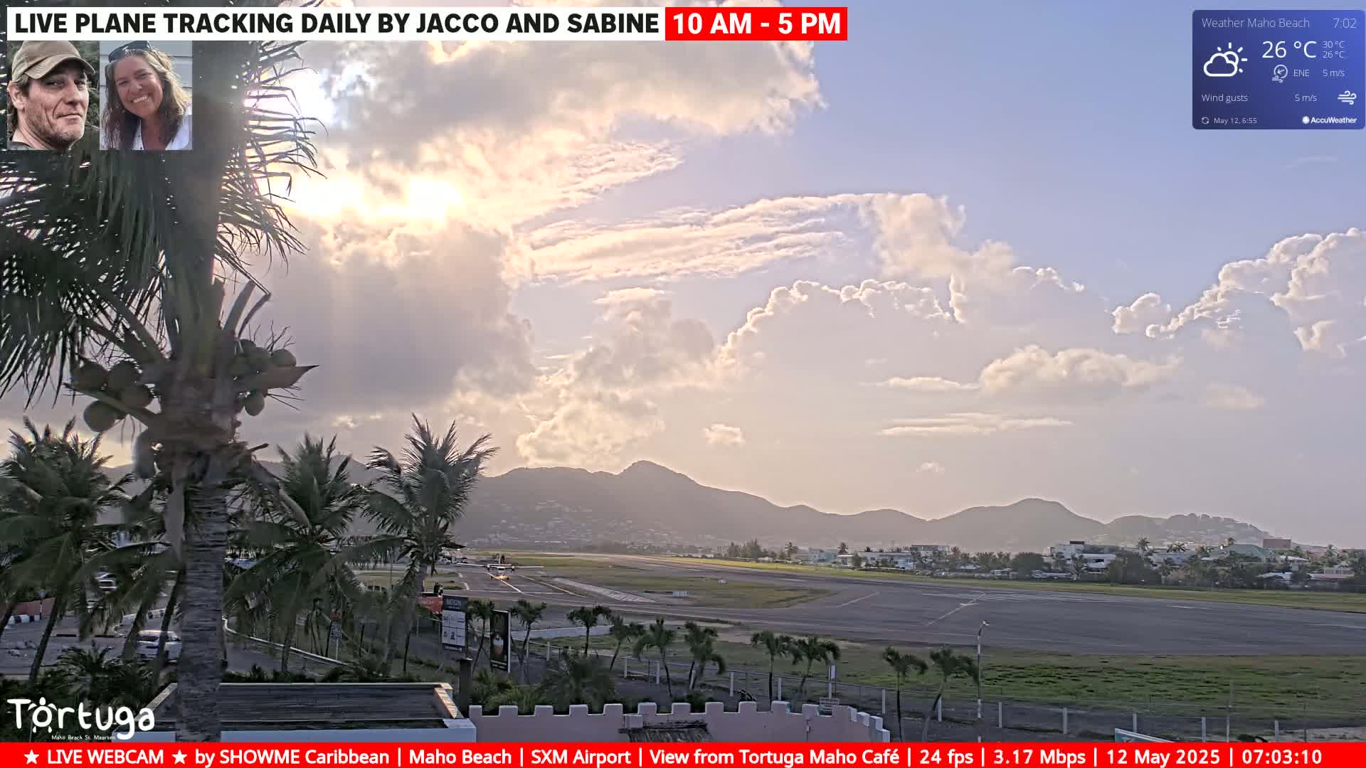 A partly cloudy sunset over an airport runway with palm trees in the foreground and mountains in the background.