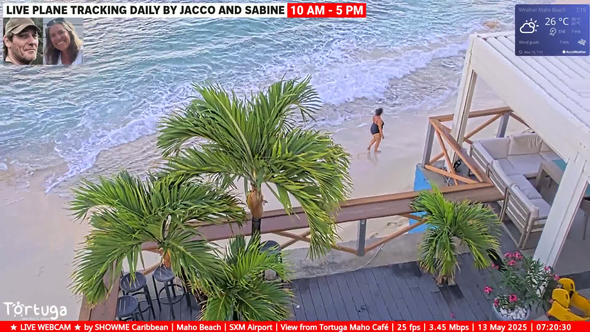 A woman walks along a sandy beach with gentle waves, near a beachfront patio with palm trees under partly sunny skies.