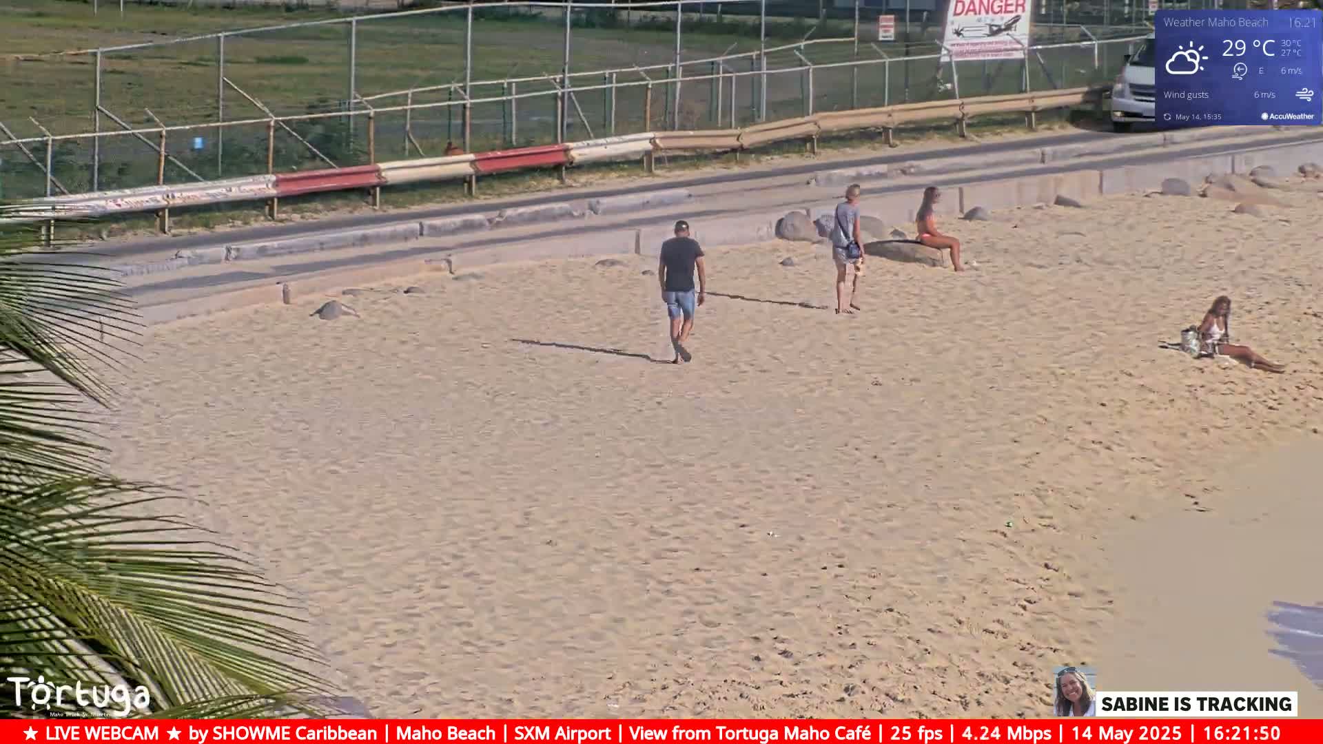 A sandy beach scene on a sunny day shows several people walking and sitting on the beach near a road and fence.