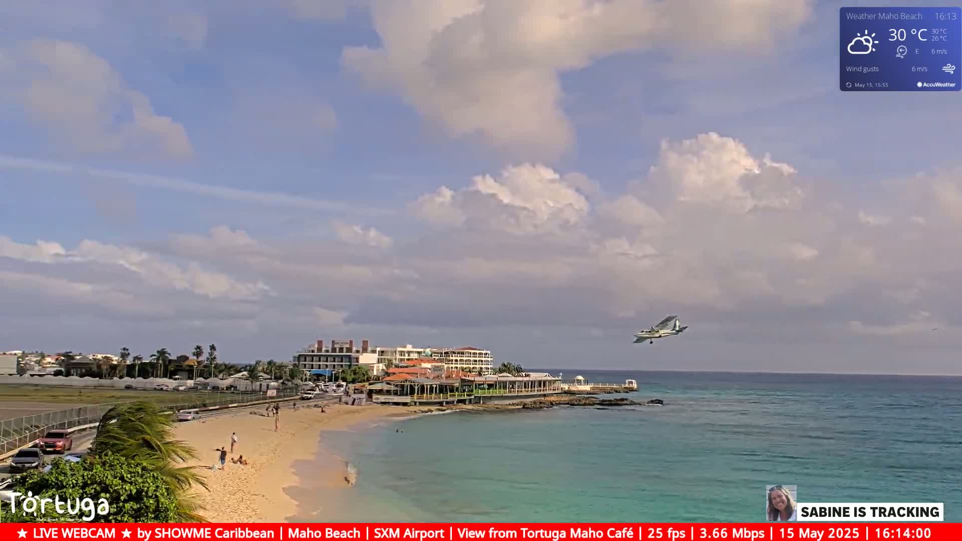 A small airplane flies over a beach with turquoise water, buildings, and a few people, under a partly cloudy sky.