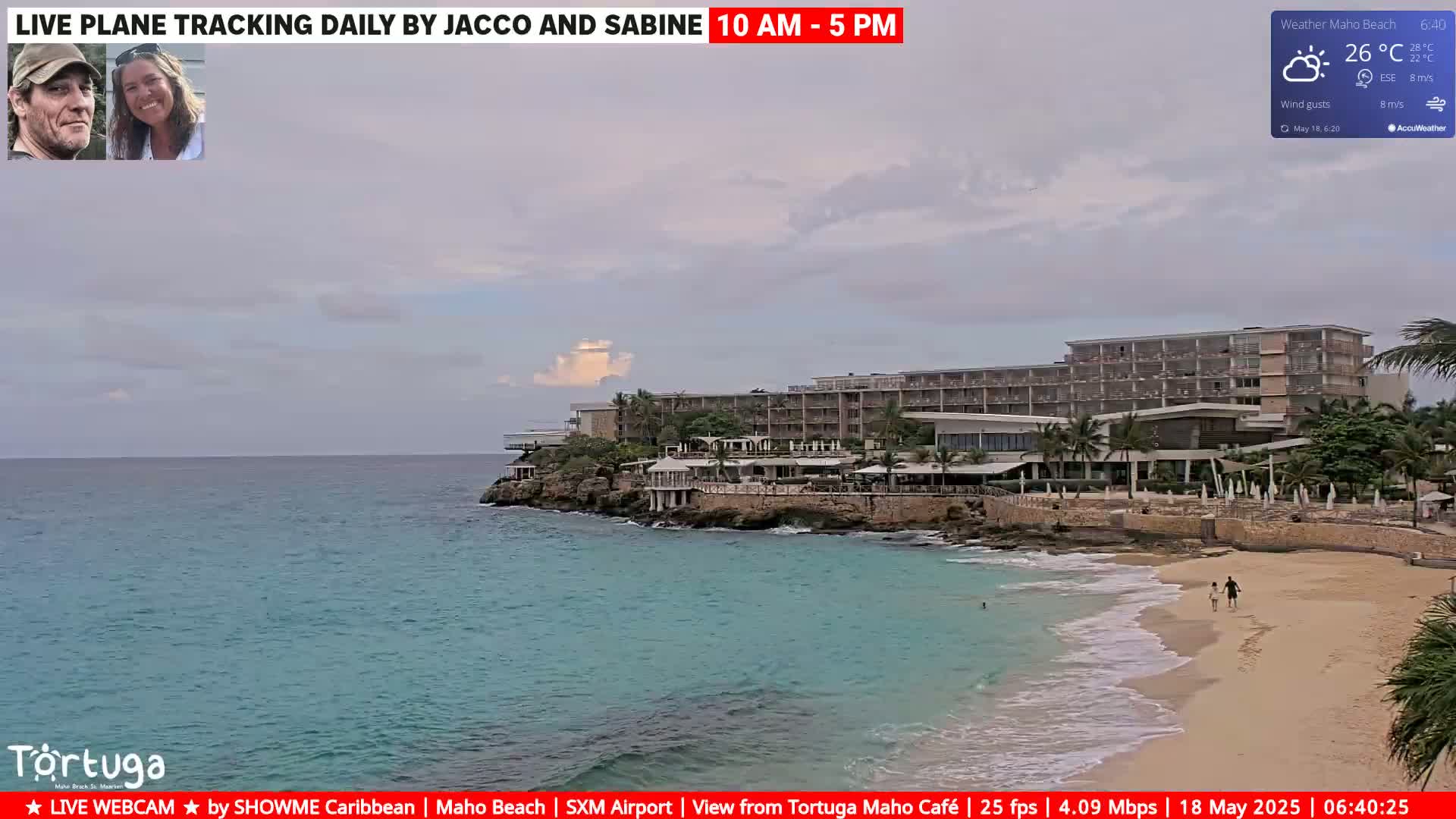 A partly cloudy day shows a beach with turquoise water, a sandy shore, a resort, and a couple walking on the beach.