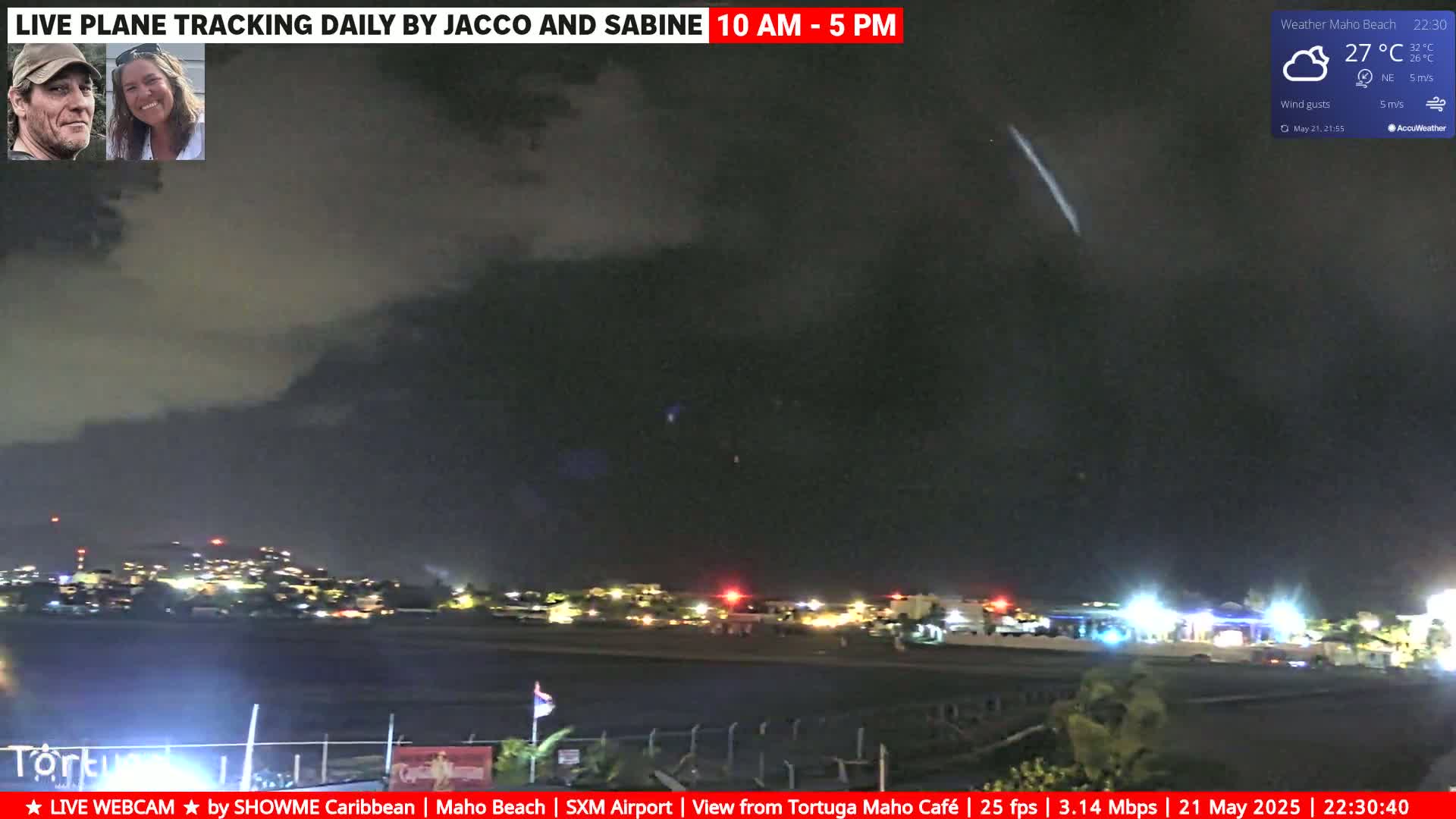 A nighttime view of an airport runway and surrounding area under a cloudy, dark sky with a single streak of light visible in the distance.