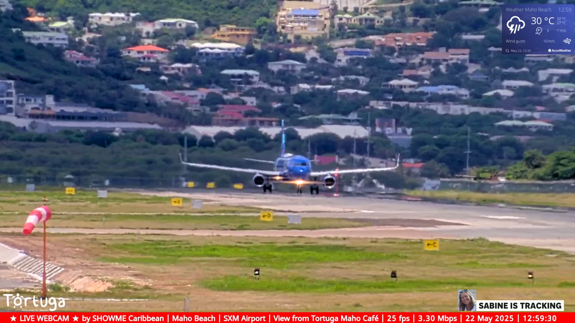 A blue and white airplane lands on a runway near a hillside town under partly cloudy skies with wind gusts.