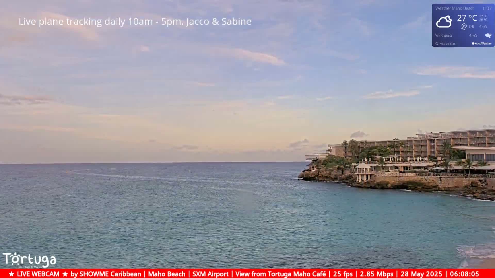 A calm ocean with a resort hotel on a rocky outcrop under a mostly clear, pale sky.