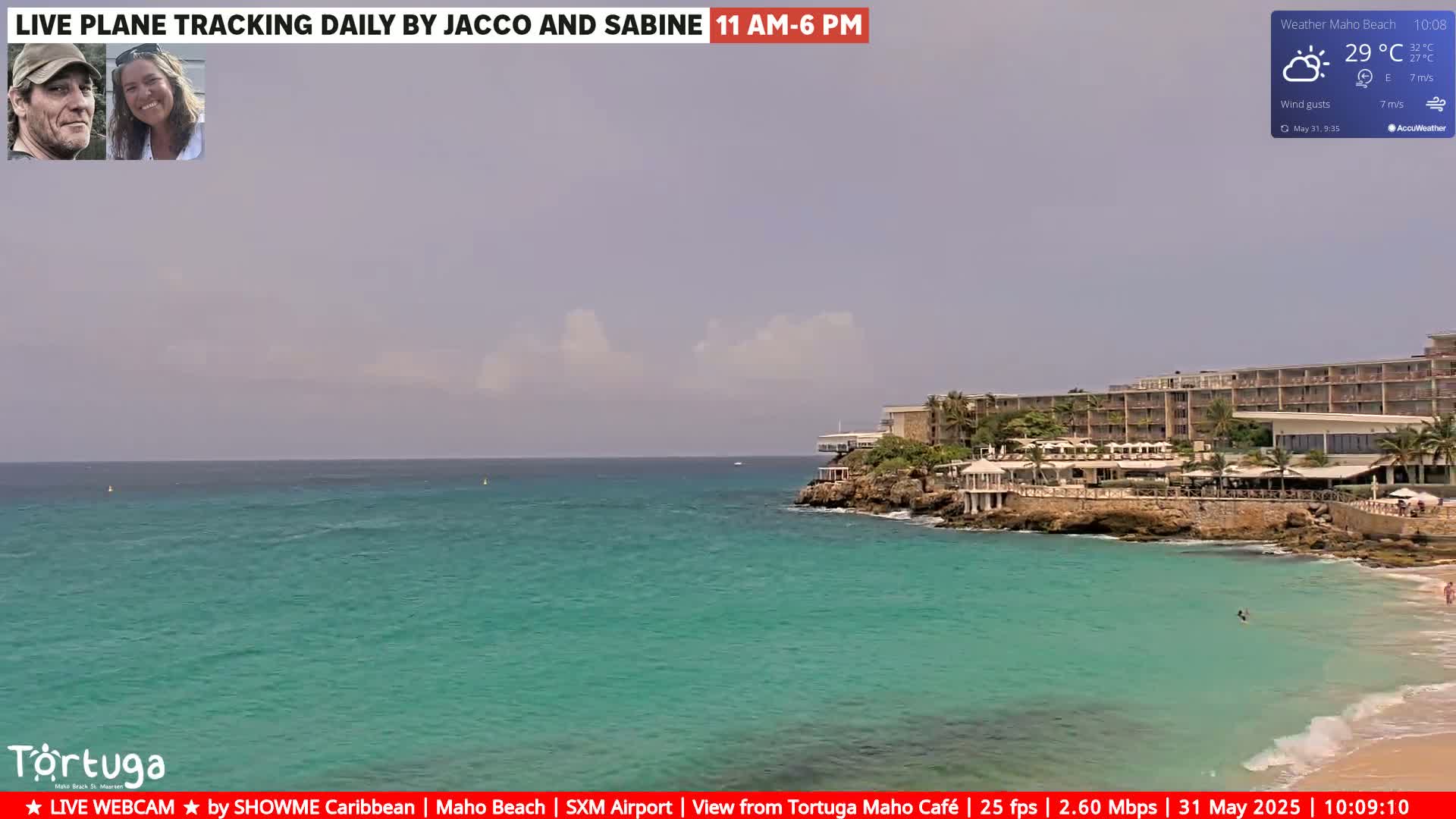 A partly cloudy day shows a turquoise ocean, a sandy beach, and a resort on a rocky outcrop.
