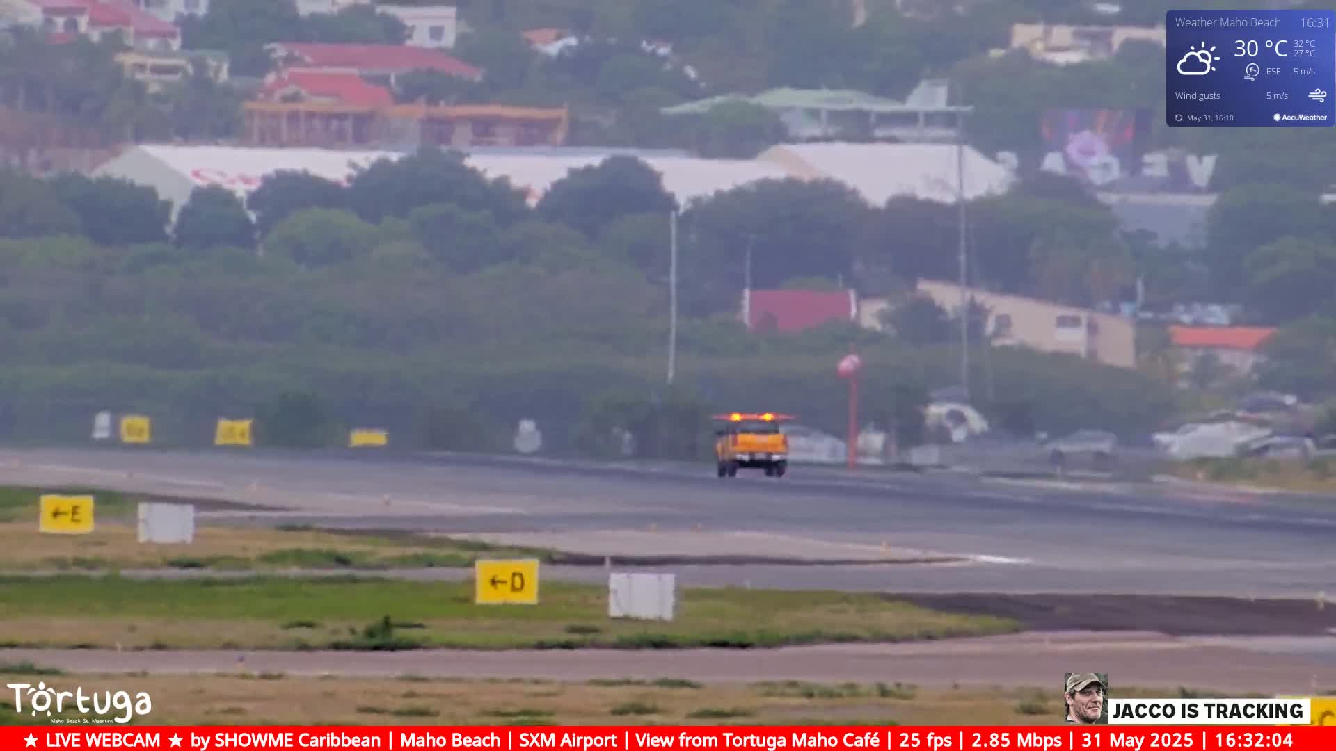A hazy view of an airport runway with a yellow vehicle, buildings in the distance, and a partly cloudy sky.
