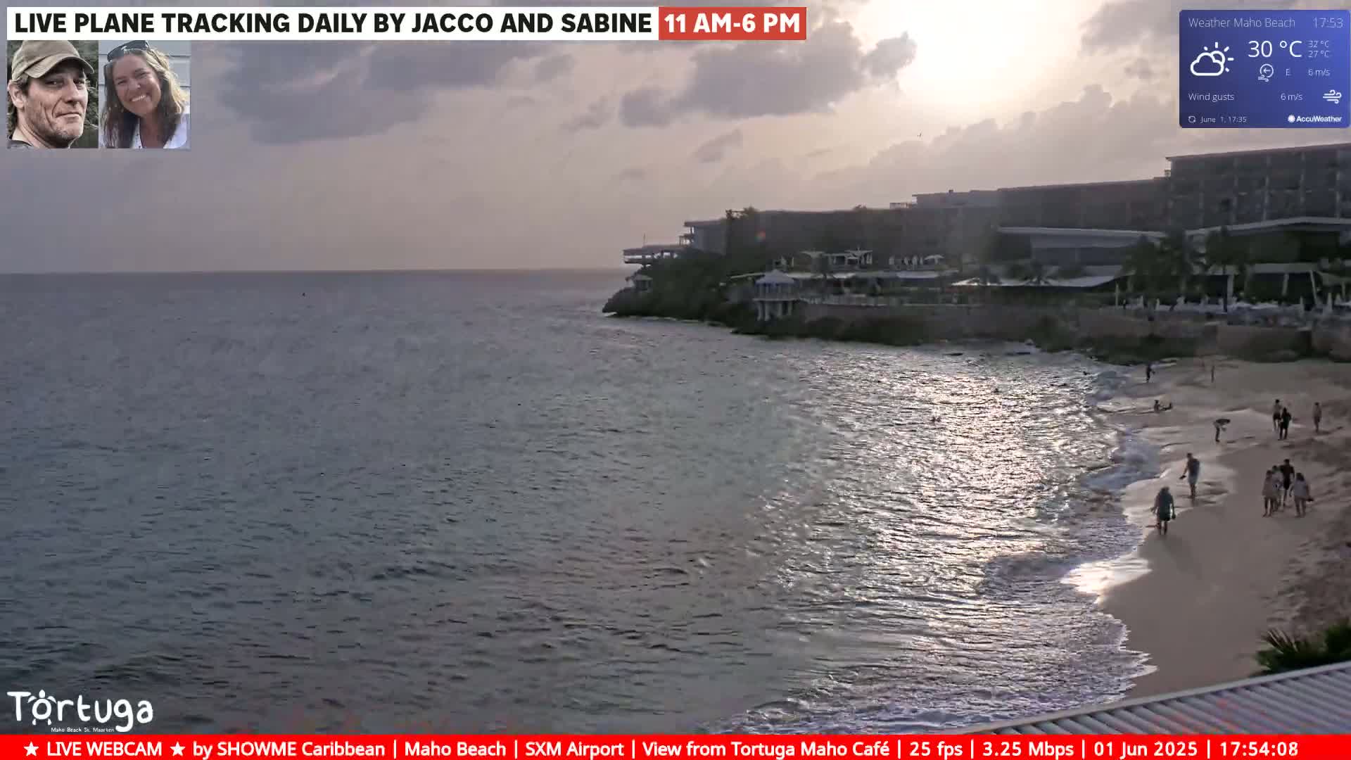 A beach with a few people walking on the sand, next to a calm ocean and a resort under a partly cloudy sunset.