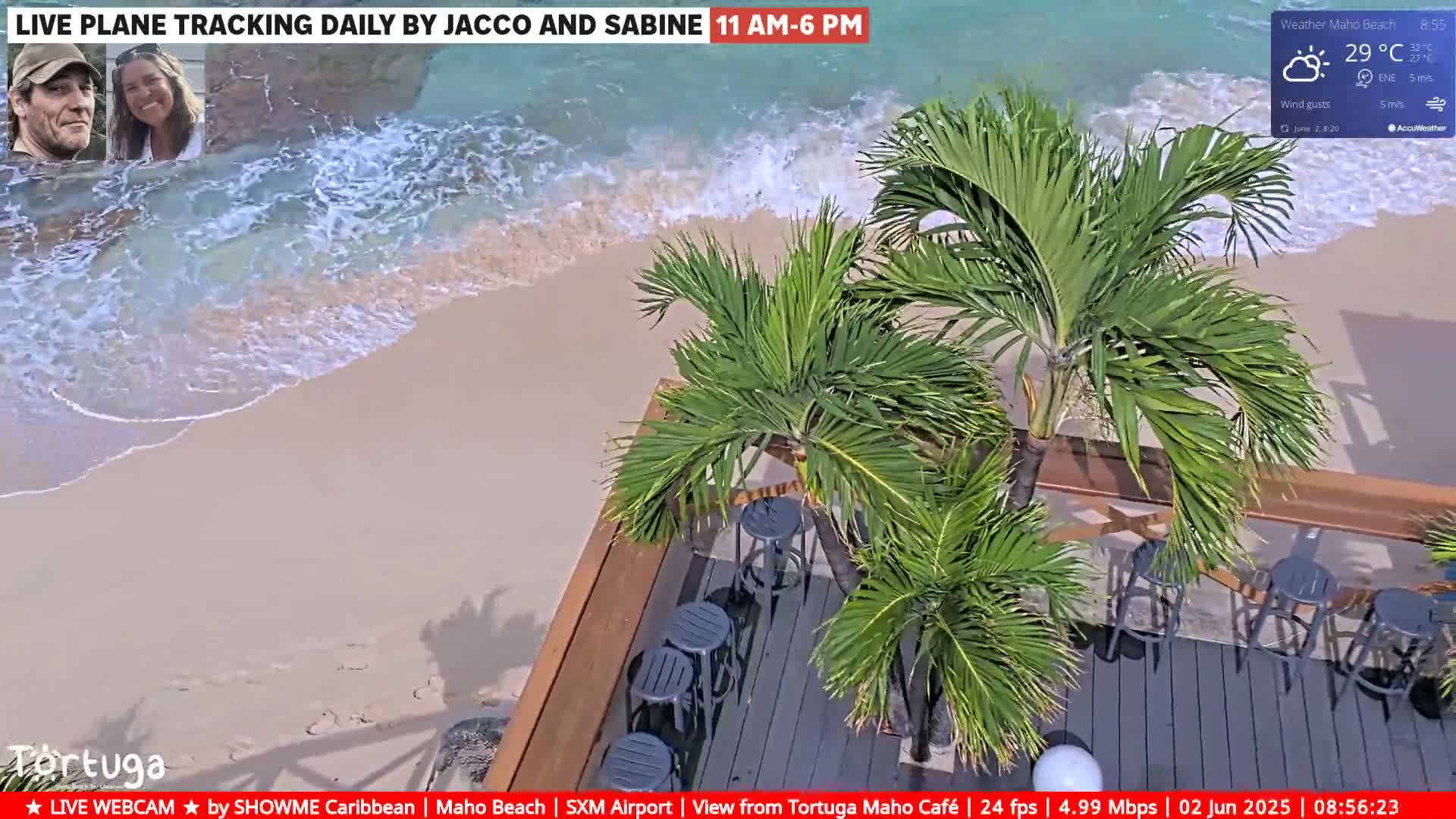 A beach with gentle waves laps a sandy shore under sunny skies, next to an outdoor patio area with palm trees and empty bar stools.