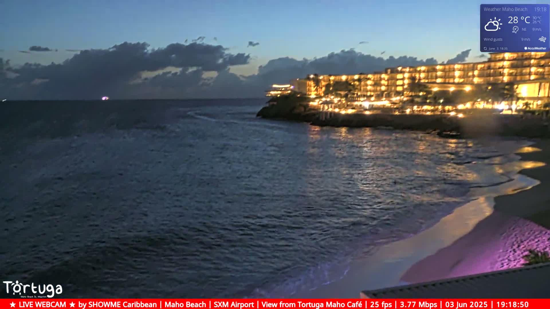 A brightly lit hotel is situated on a dark, rocky coast overlooking a calm ocean at dusk under a partly cloudy sky.