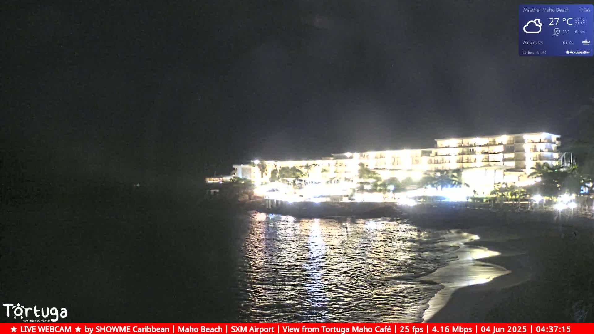 A brightly lit hotel sits on a dark beach at night, with calm water reflecting its lights.