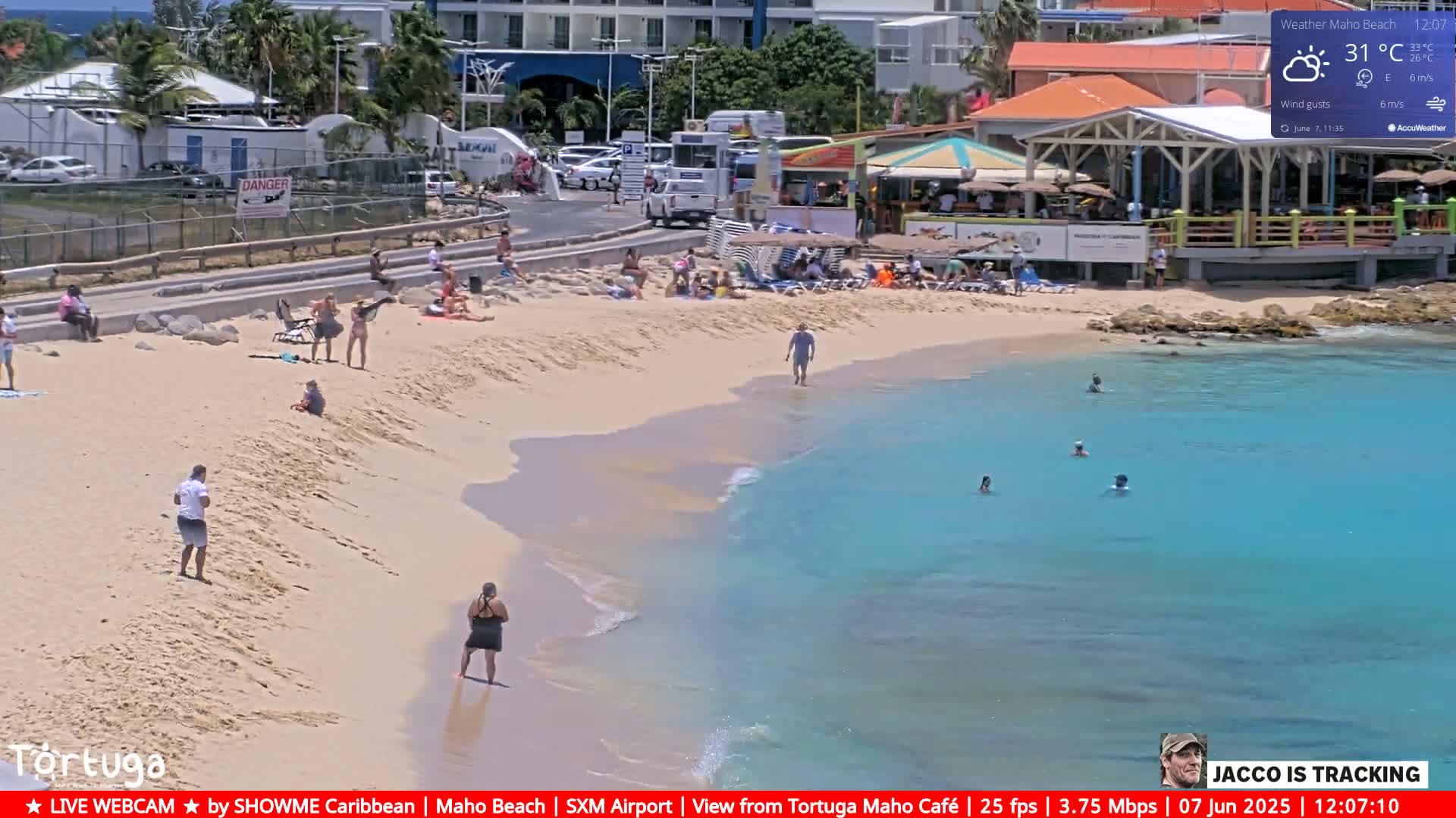 A sandy beach with turquoise water and numerous people sunbathing, swimming, and relaxing near beachside buildings under partly sunny skies.