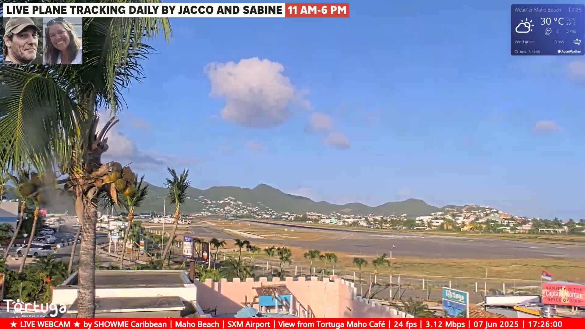 A sunny day shows a palm tree-lined airport runway and surrounding buildings with a partly cloudy sky and distant mountains.