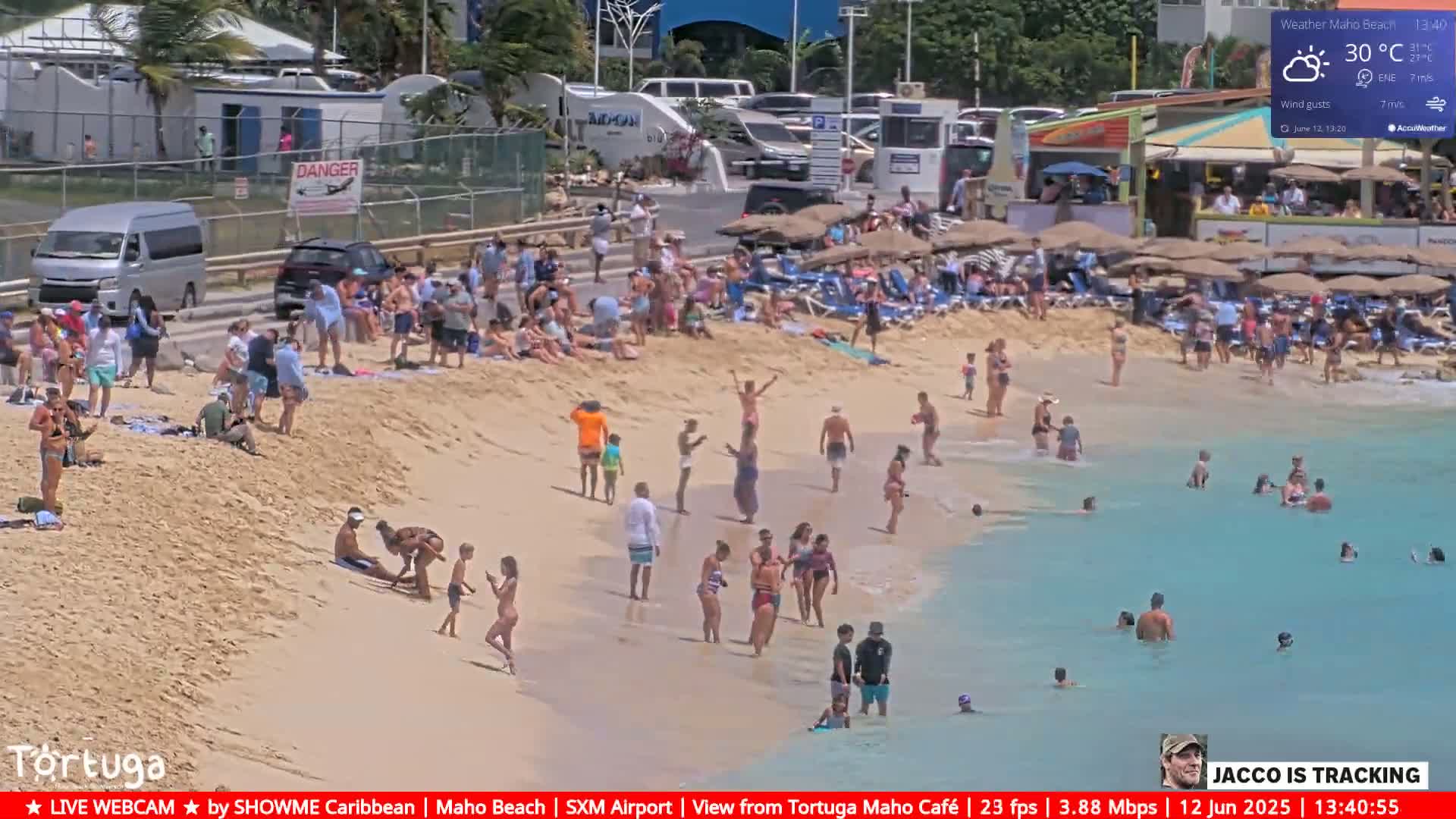 A sunny beach scene shows many people relaxing, swimming, and sunbathing on a sandy beach with turquoise water, under a partly cloudy sky.