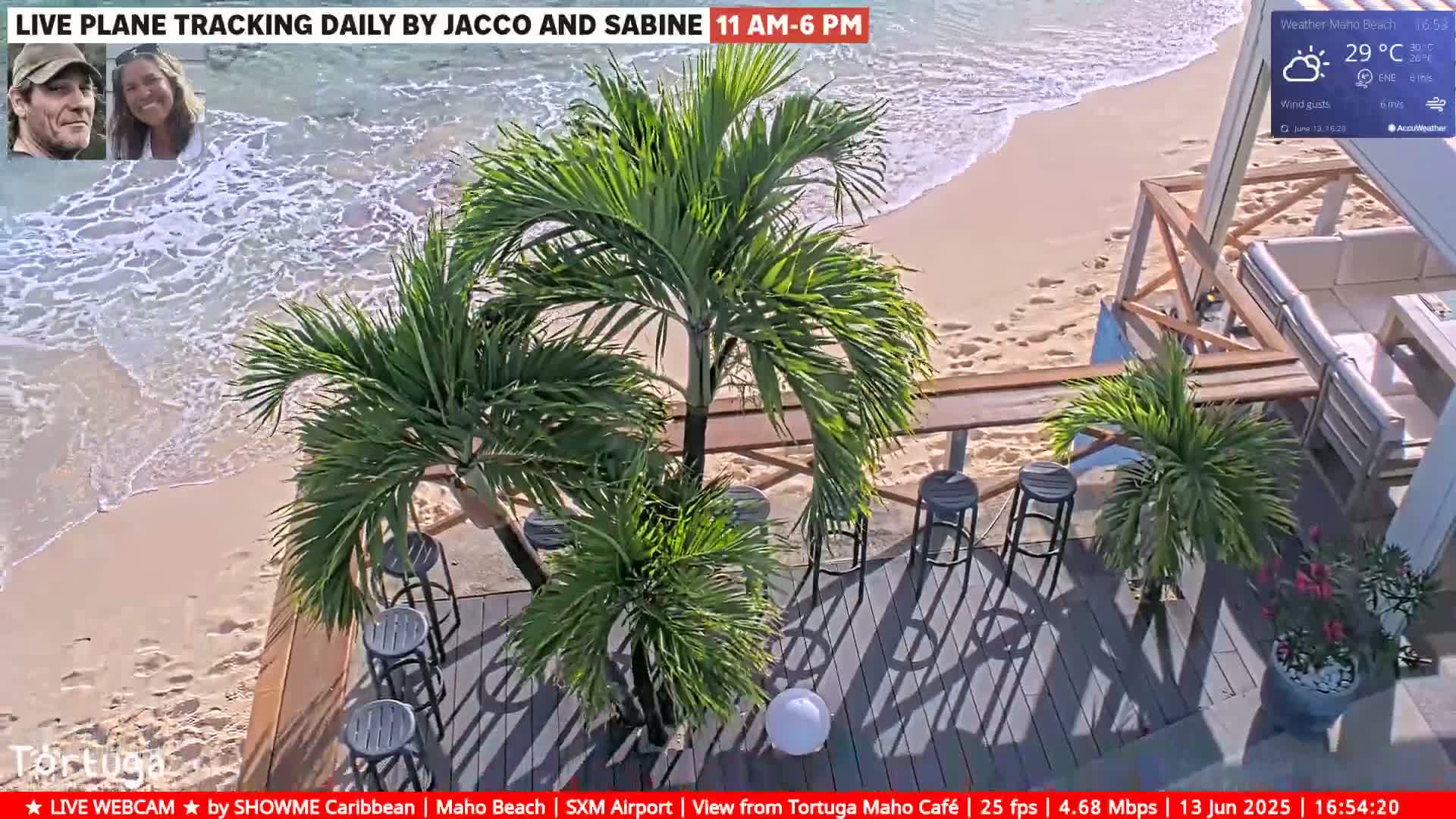 An outdoor beachside cafe with palm trees and gray bar stools sits on a wooden deck overlooking a sandy beach with gentle waves under sunny skies.