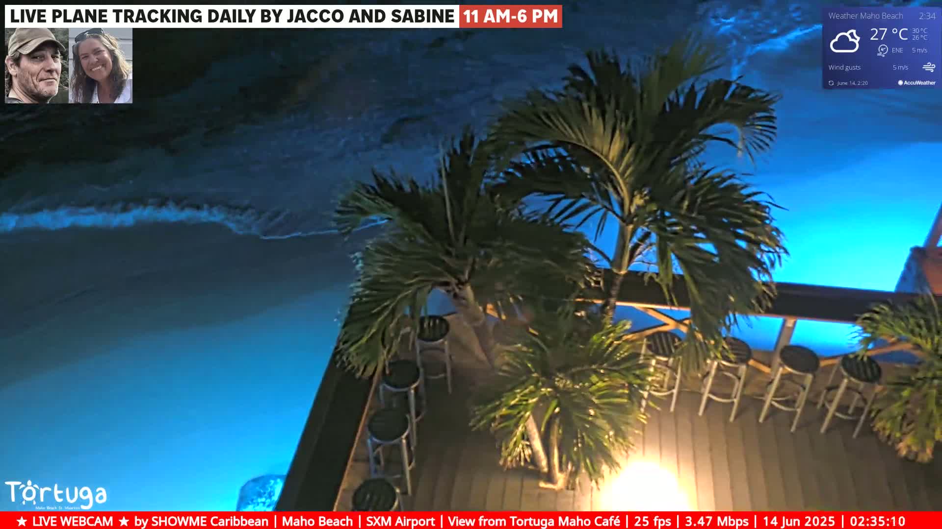 An outdoor beachside cafe with palm trees and gray bar stools sits on a wooden deck overlooking a sandy beach with gentle waves under sunny skies.