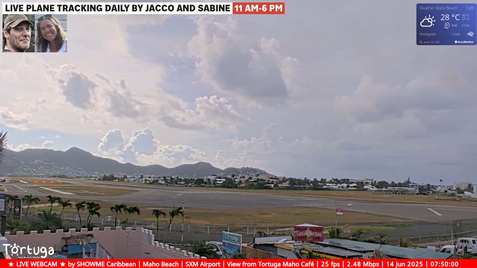 A partly cloudy daytime view of an airport runway, surrounded by buildings and palm trees, with mountains in the background.
