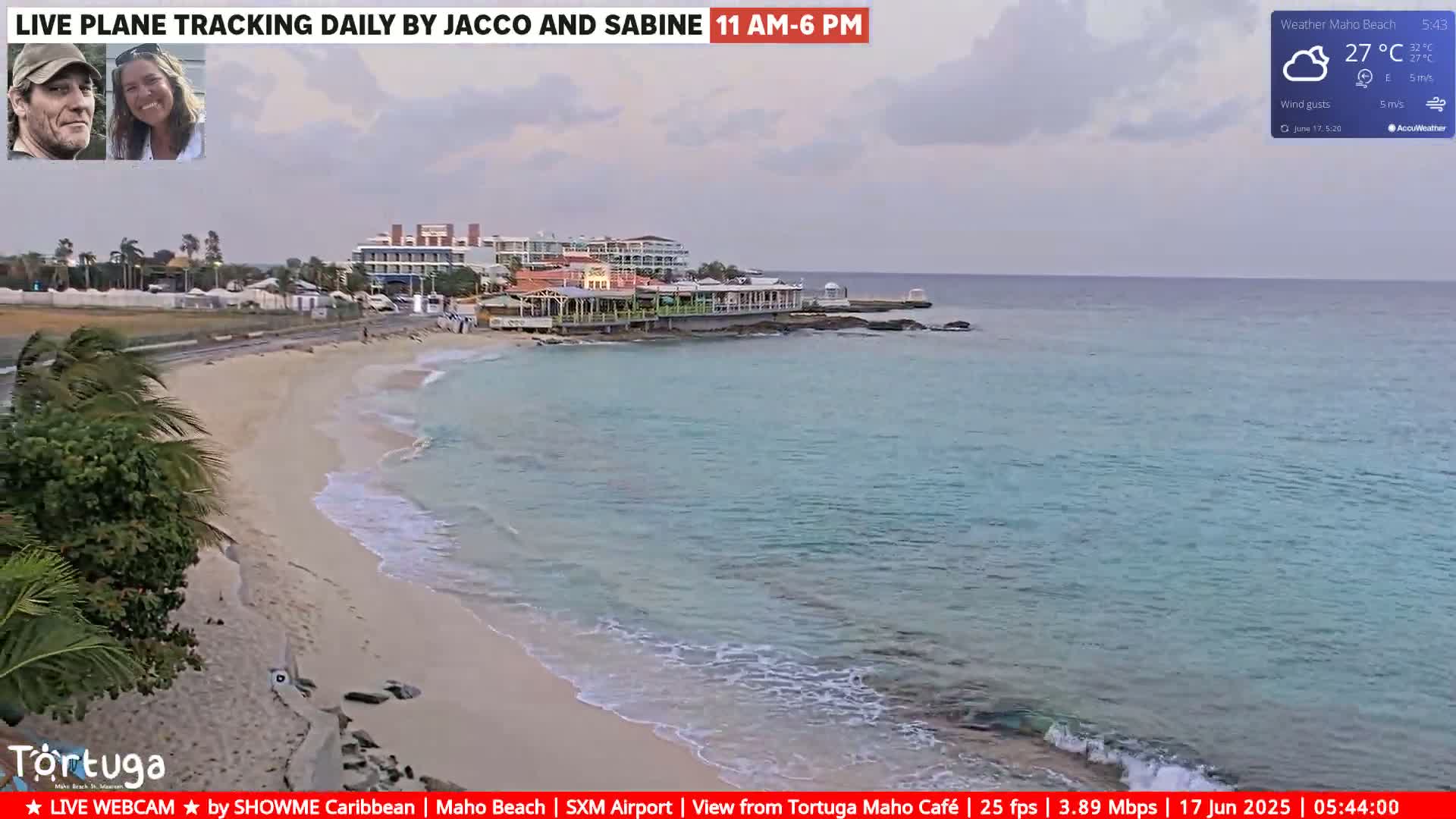 A beach with light-colored sand and calm, light-blue water is seen under a partly cloudy sky, with buildings and a restaurant visible in the background.