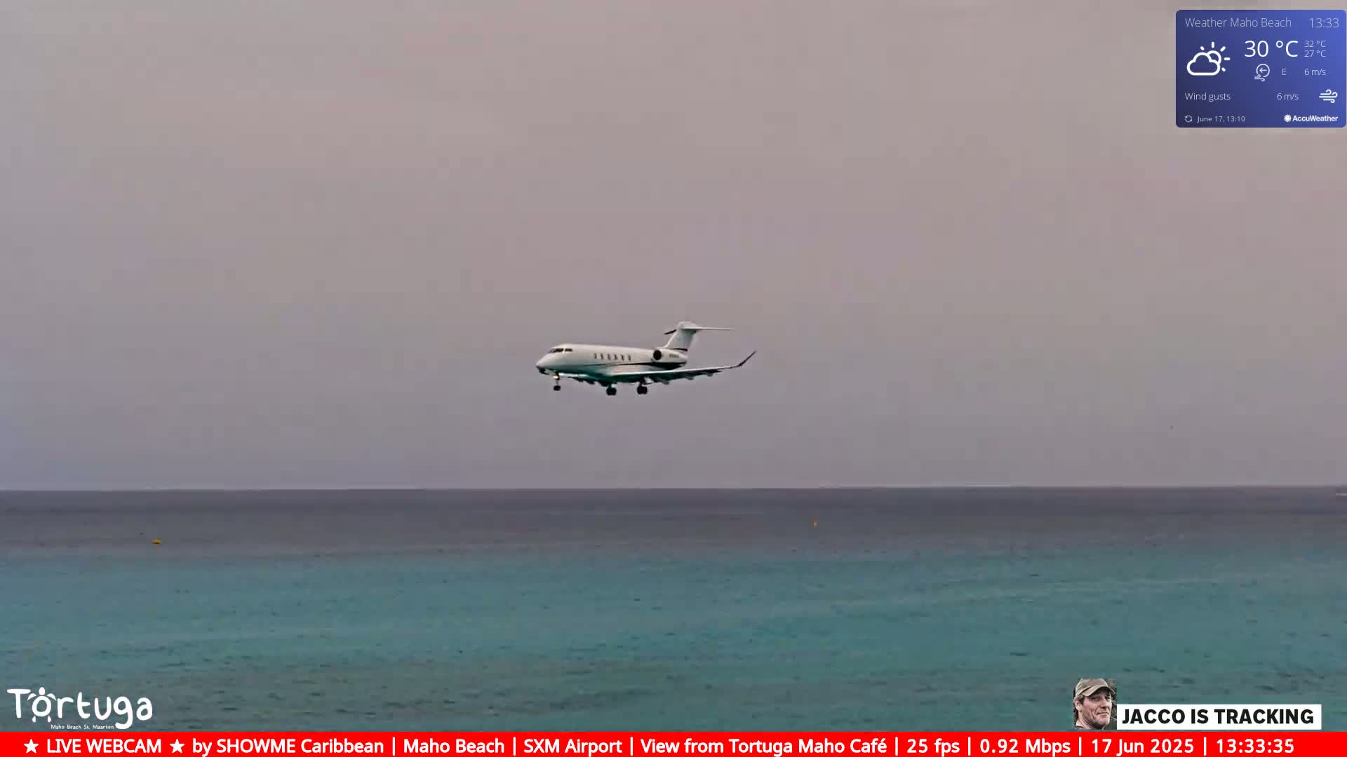 A white private jet flies low over a calm, turquoise ocean under a hazy, light gray sky.