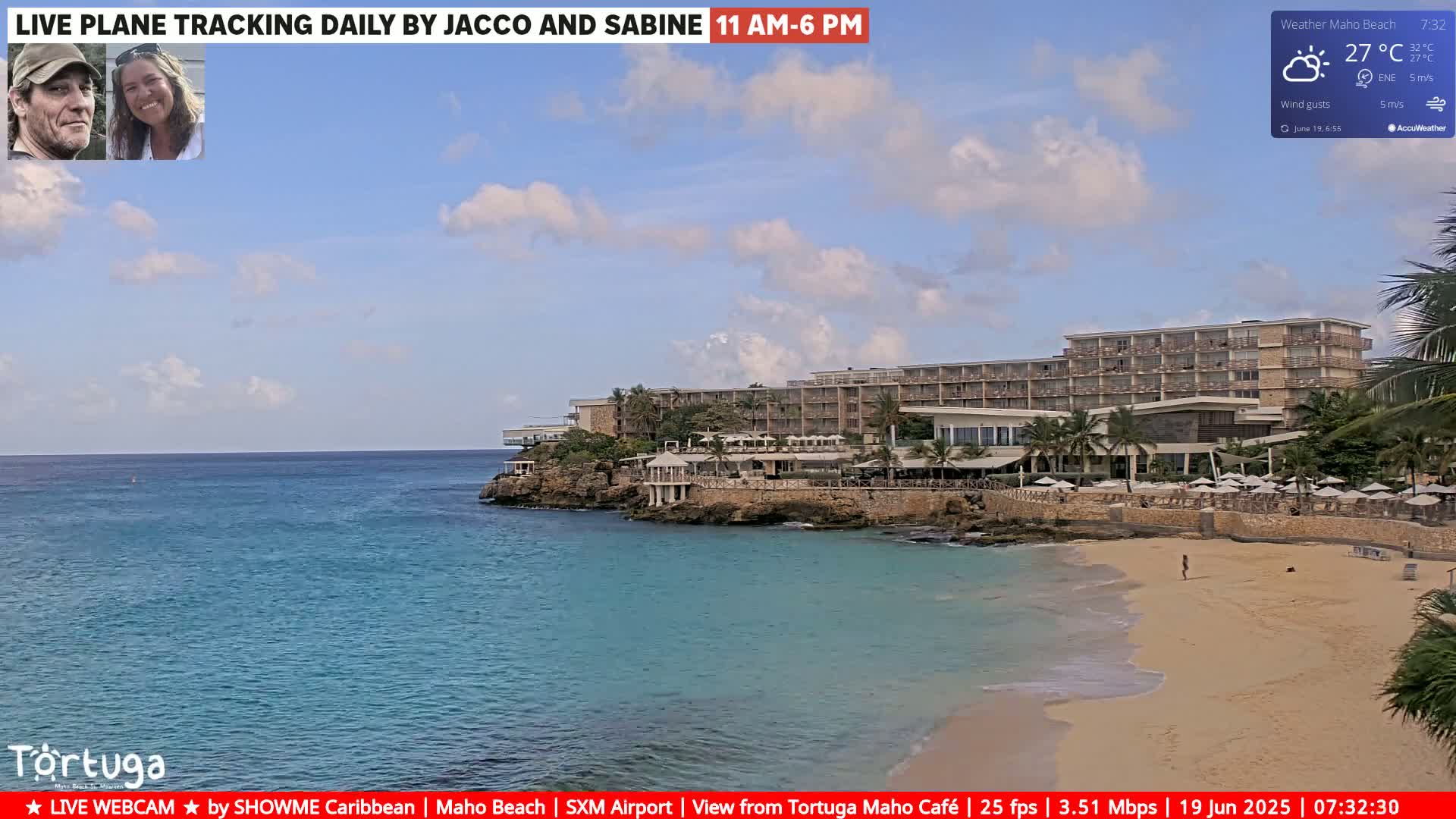 A partly cloudy sunny day shows a tranquil beach scene with a large hotel on a rocky outcrop, calm turquoise water, and a sandy beach.