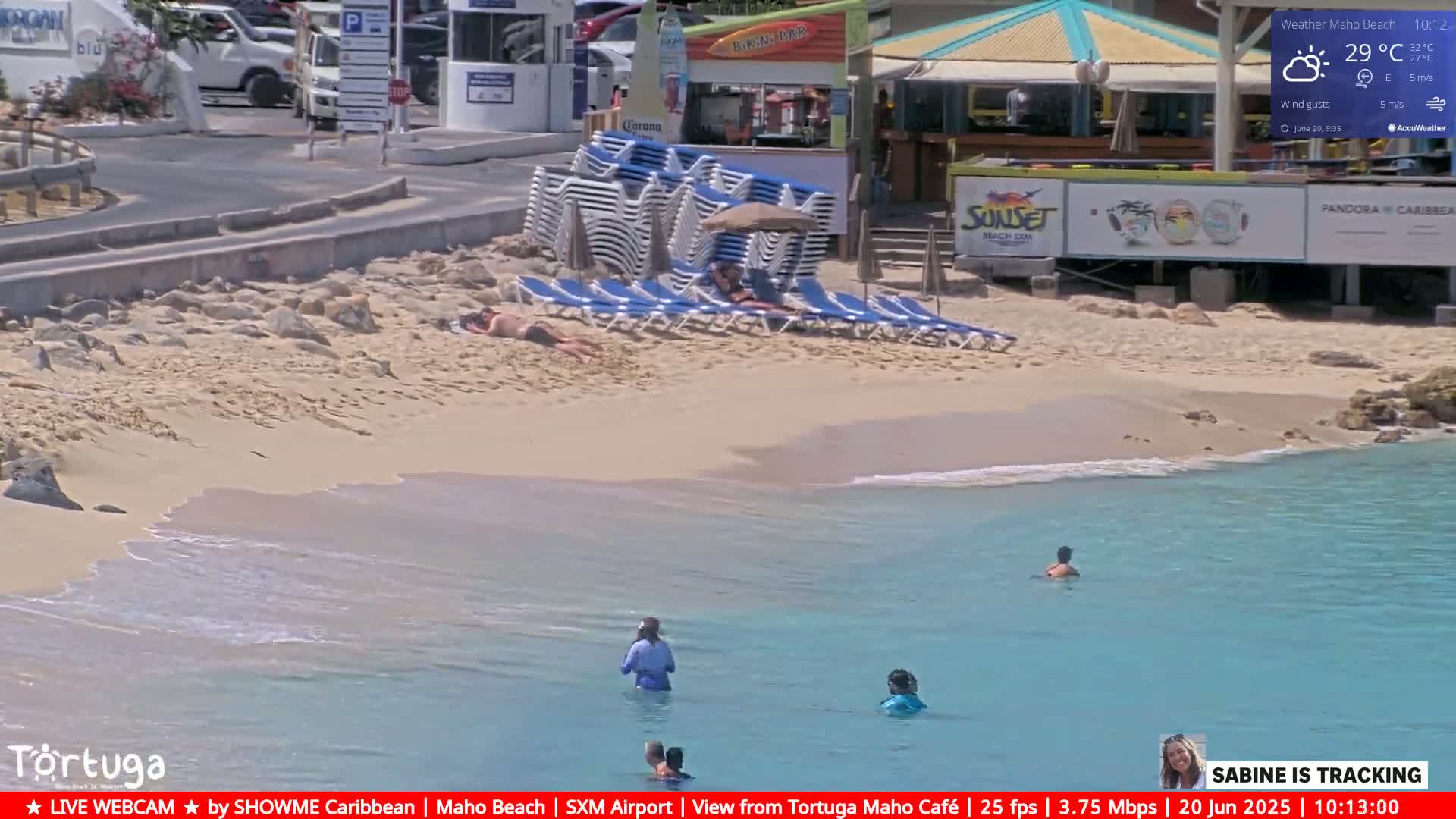 A sunny beach with calm, turquoise water shows people swimming and sunbathing near beach chairs and umbrellas stacked near a beachfront bar.