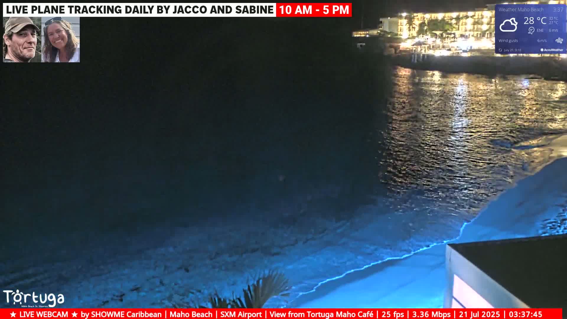 A nighttime view of a beach with calm, blue-lit water under a clear sky, showing a building and its lights reflected on the water.